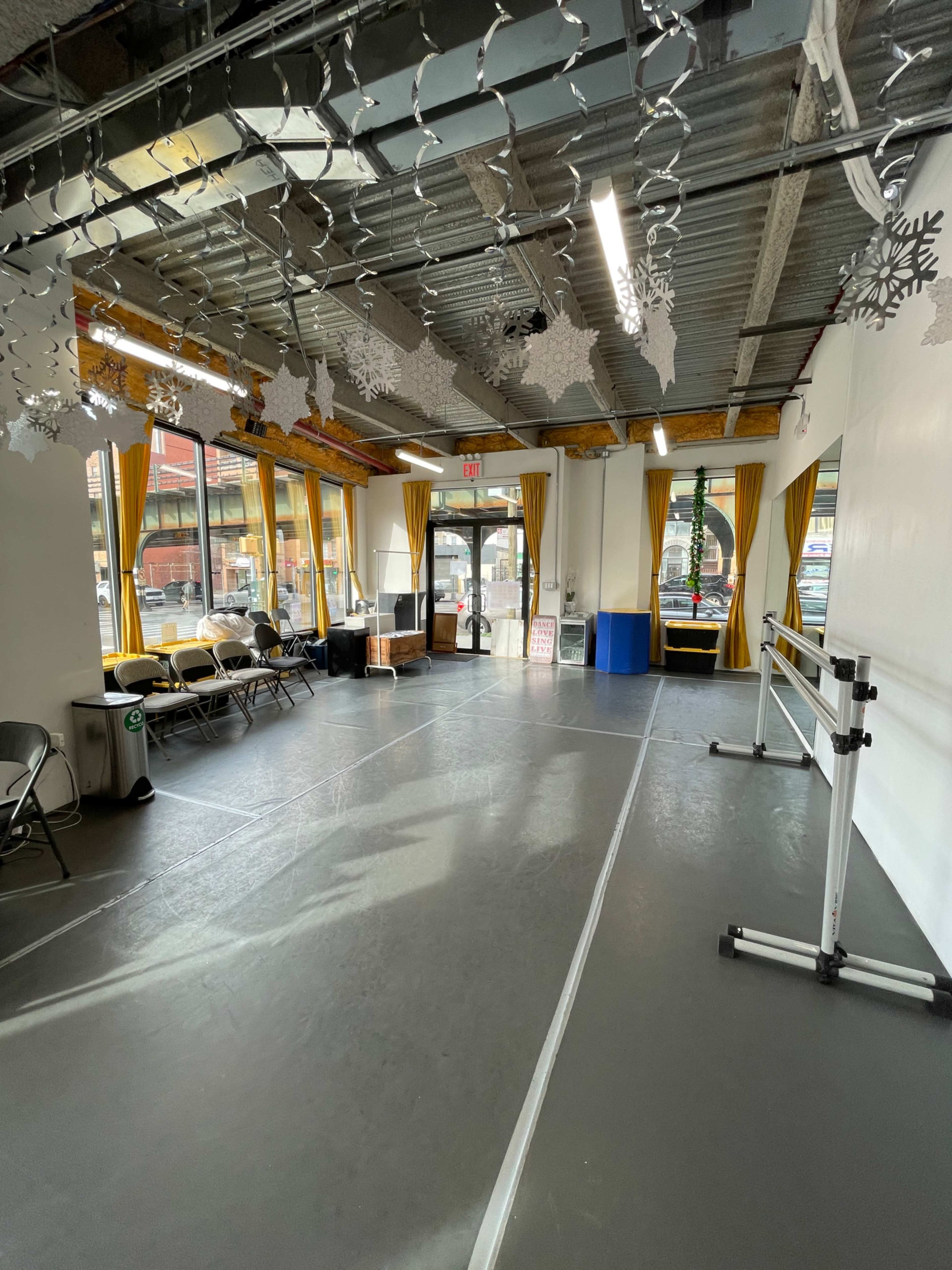 A bright room with a gray floor, large windows, and chairs arranged along the walls, featuring ballet barres on one side and winter decorations hanging from the ceiling.