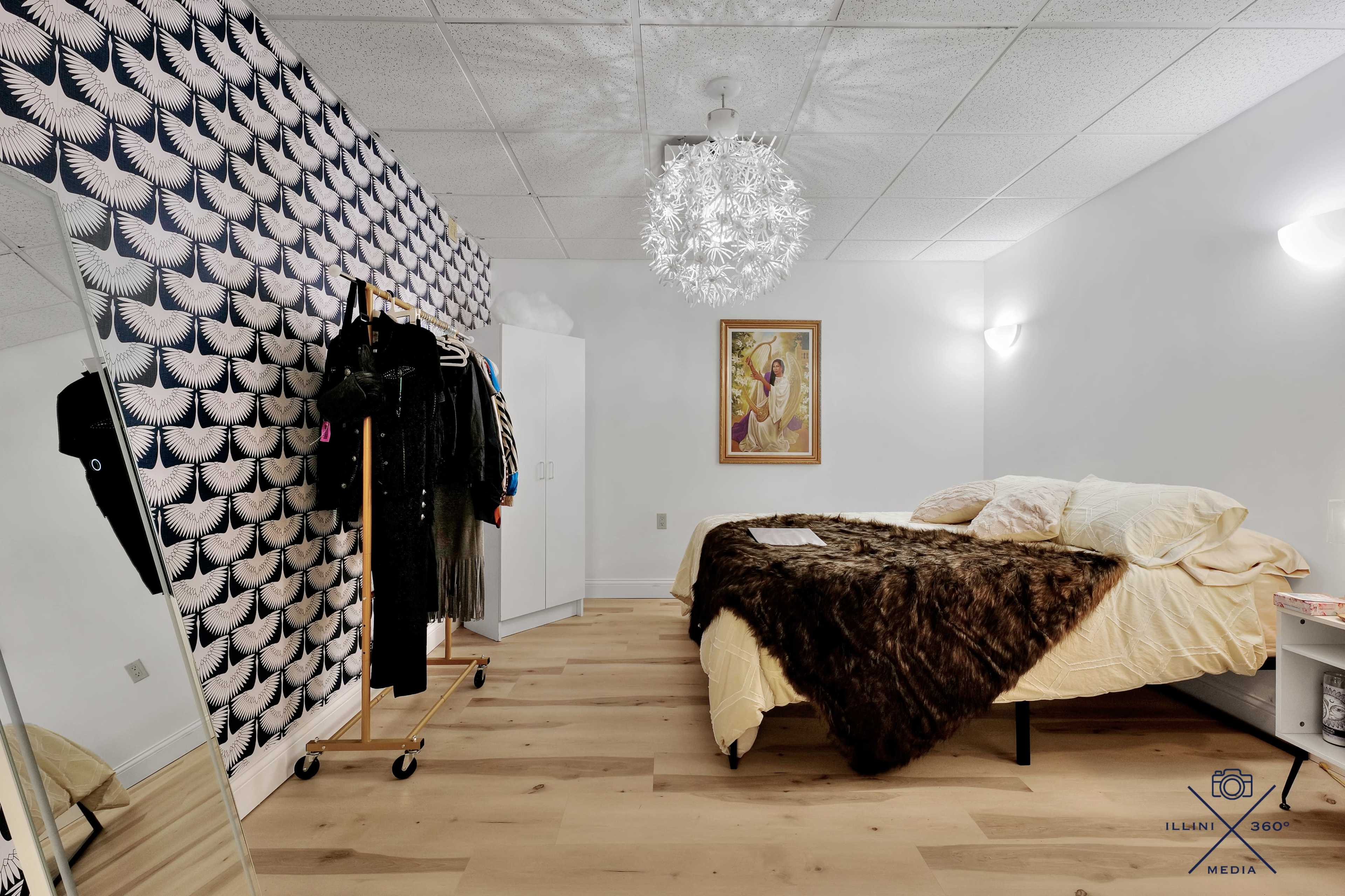 The room features a bed with a brown blanket, a mirrored wardrobe, and a patterned wall with hanging clothing on a rack.