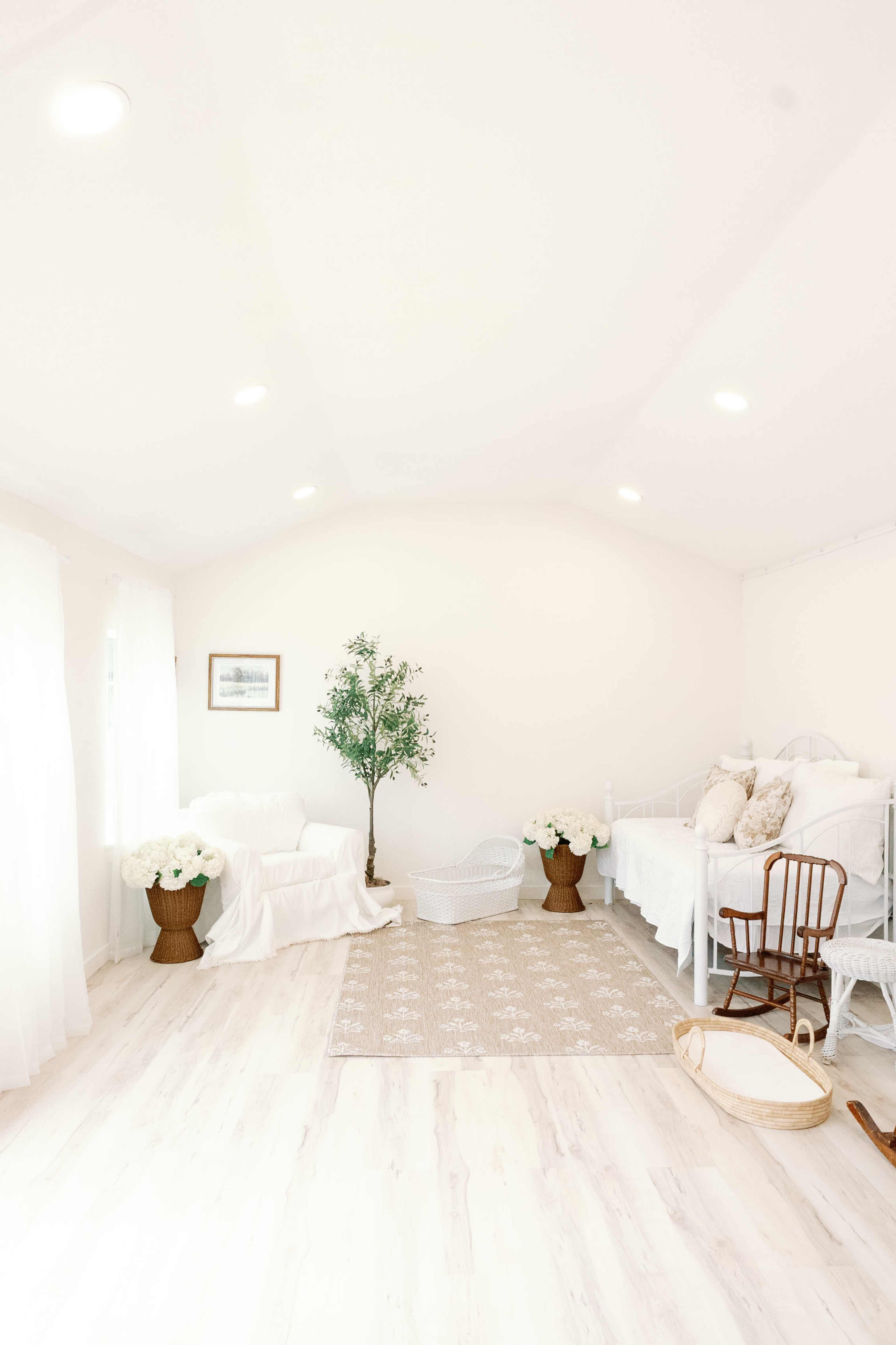 The image shows a bright, minimalistic room with a cozy seating area featuring a white sofa, a small tree, and two beds adorned with light-colored linens, complemented by decorative floral arrangements.