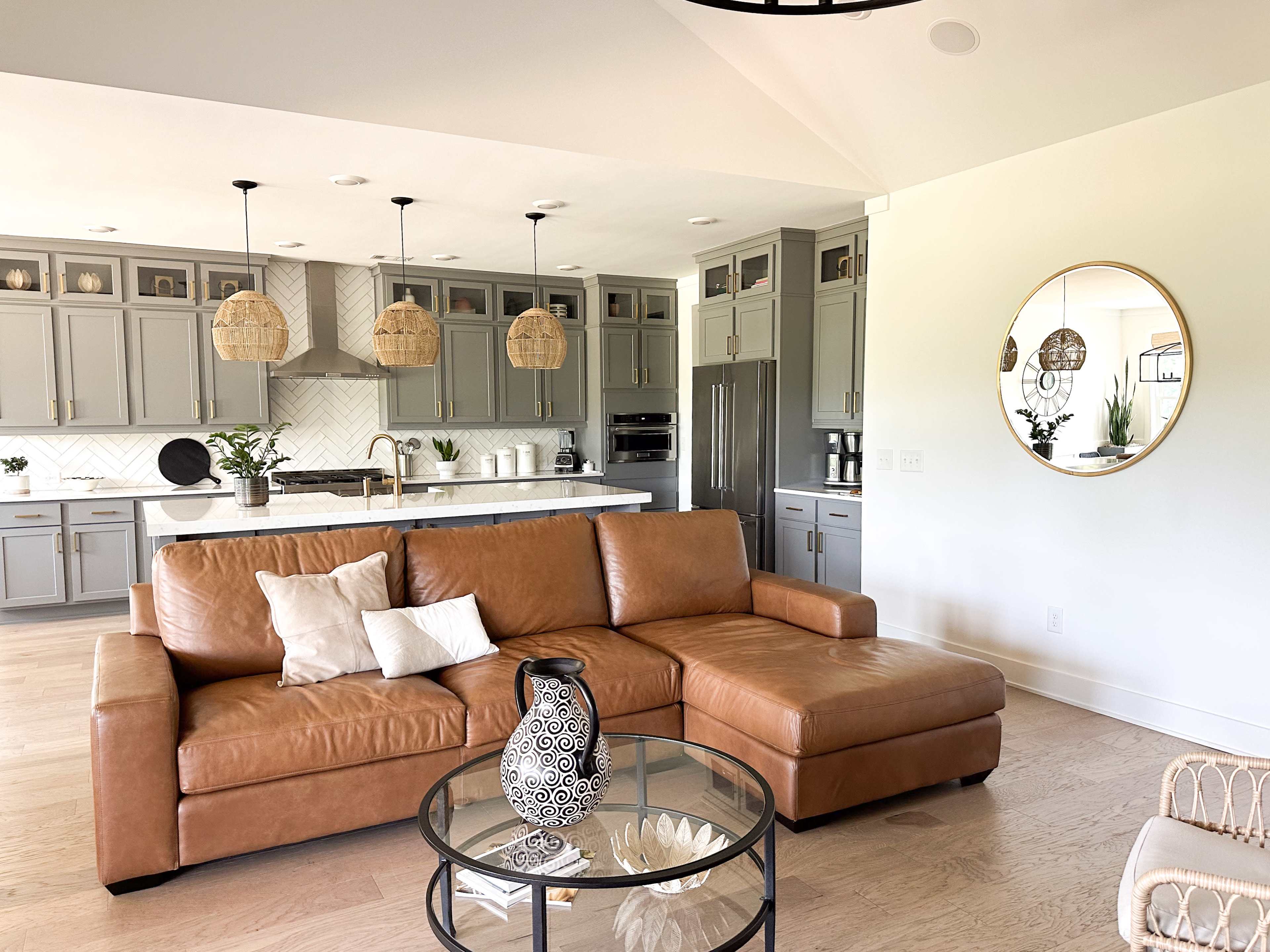 A modern living room features a brown leather sectional sofa, a glass coffee table, and a kitchen in the background with gray cabinets and pendant lights.