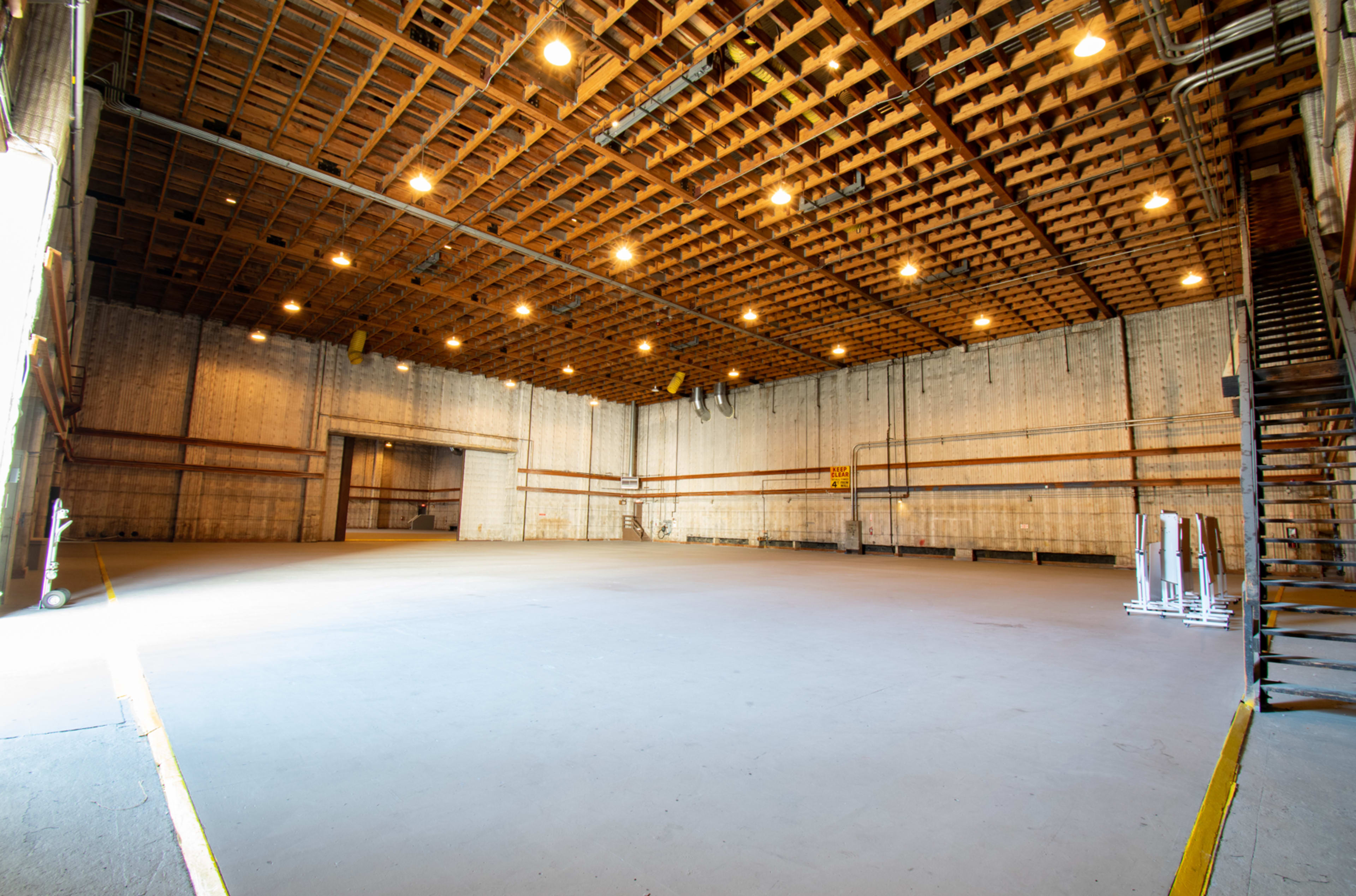 The image shows a large, empty warehouse with high wooden beams and overhead lights illuminating the concrete floor.