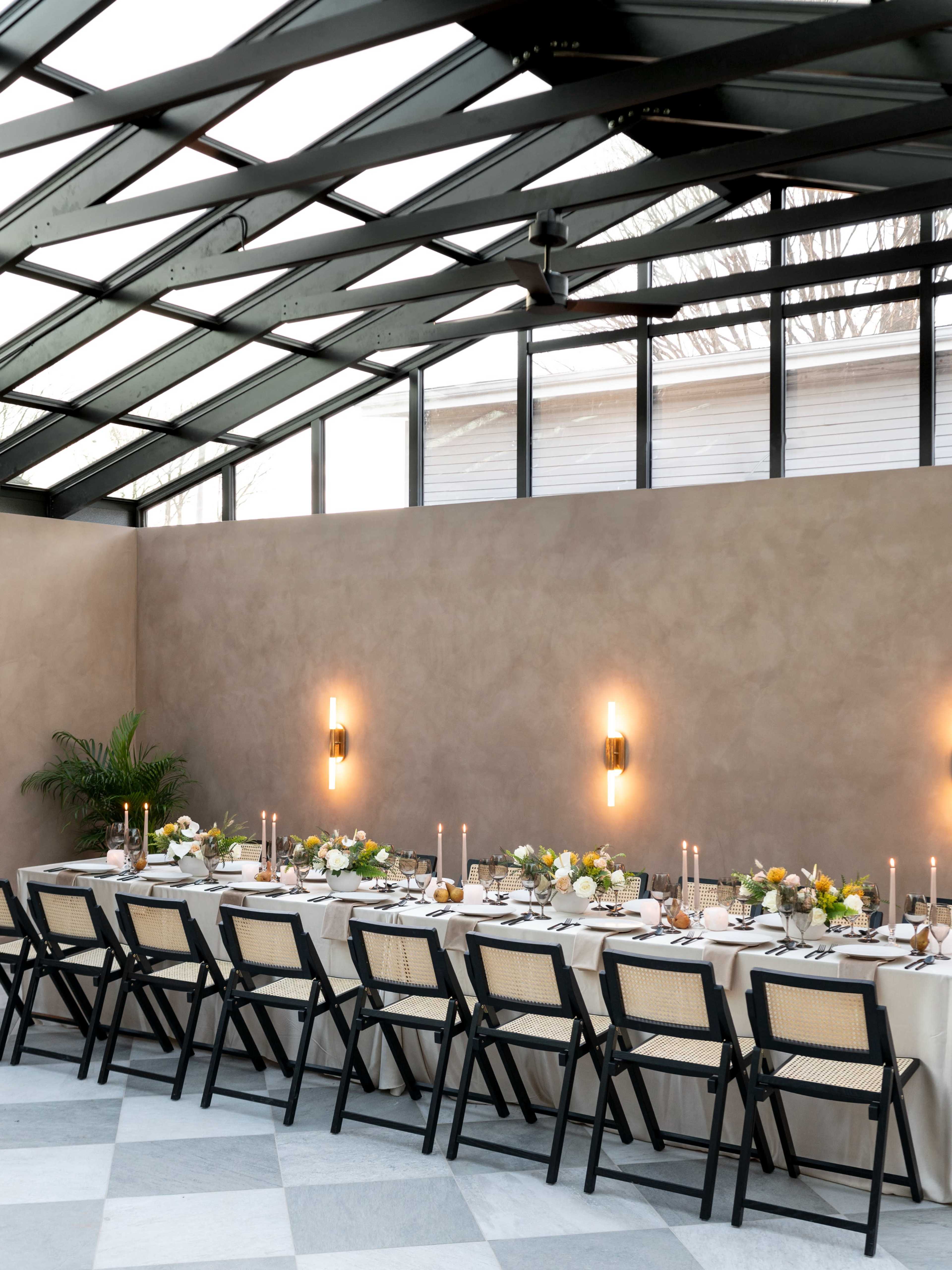 A long dining table is set with candles and flowers inside a glass-walled greenhouse.