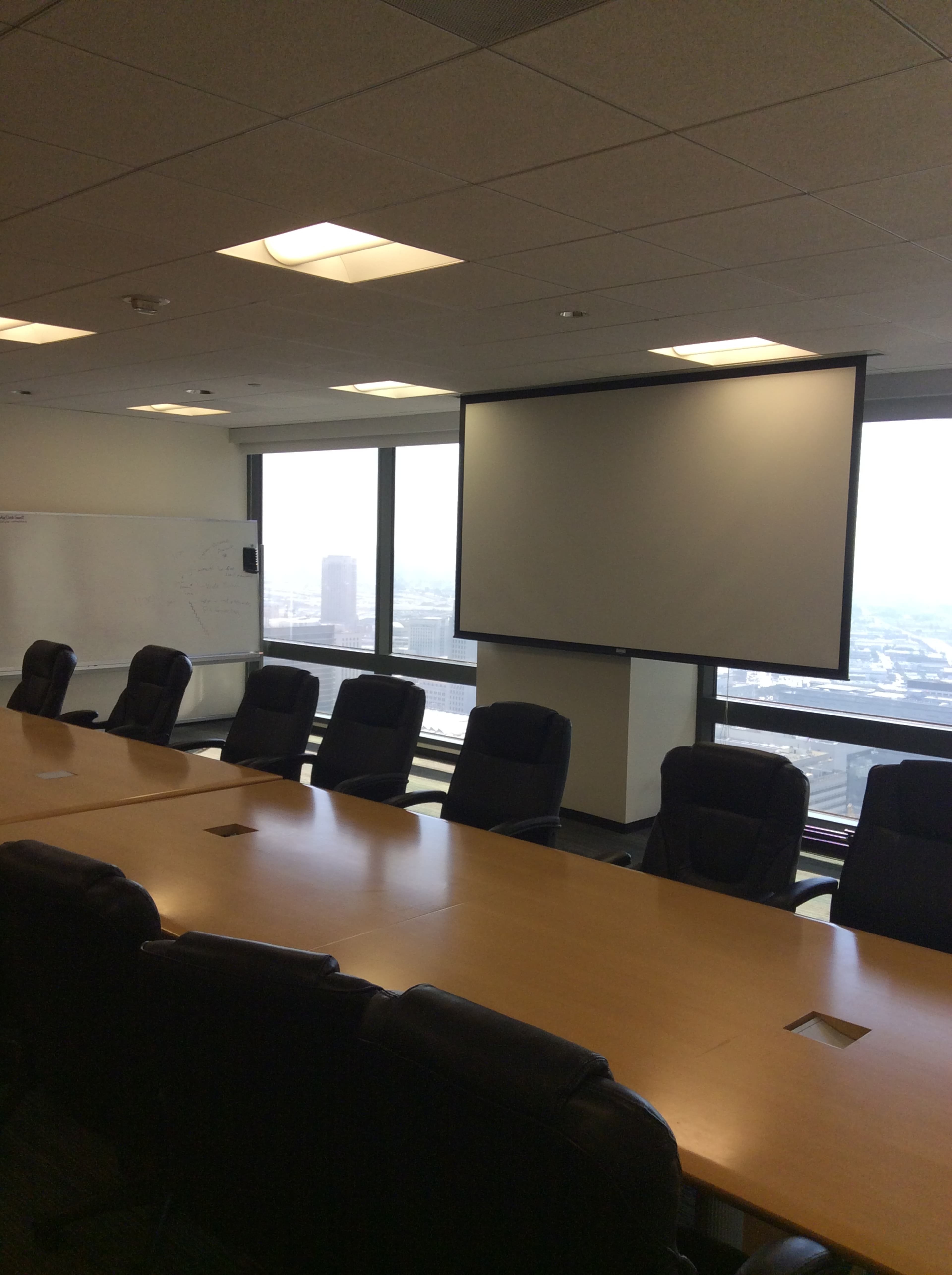A modern conference room features a large table surrounded by office chairs, with a projection screen mounted on the wall and windows providing a view of the city.