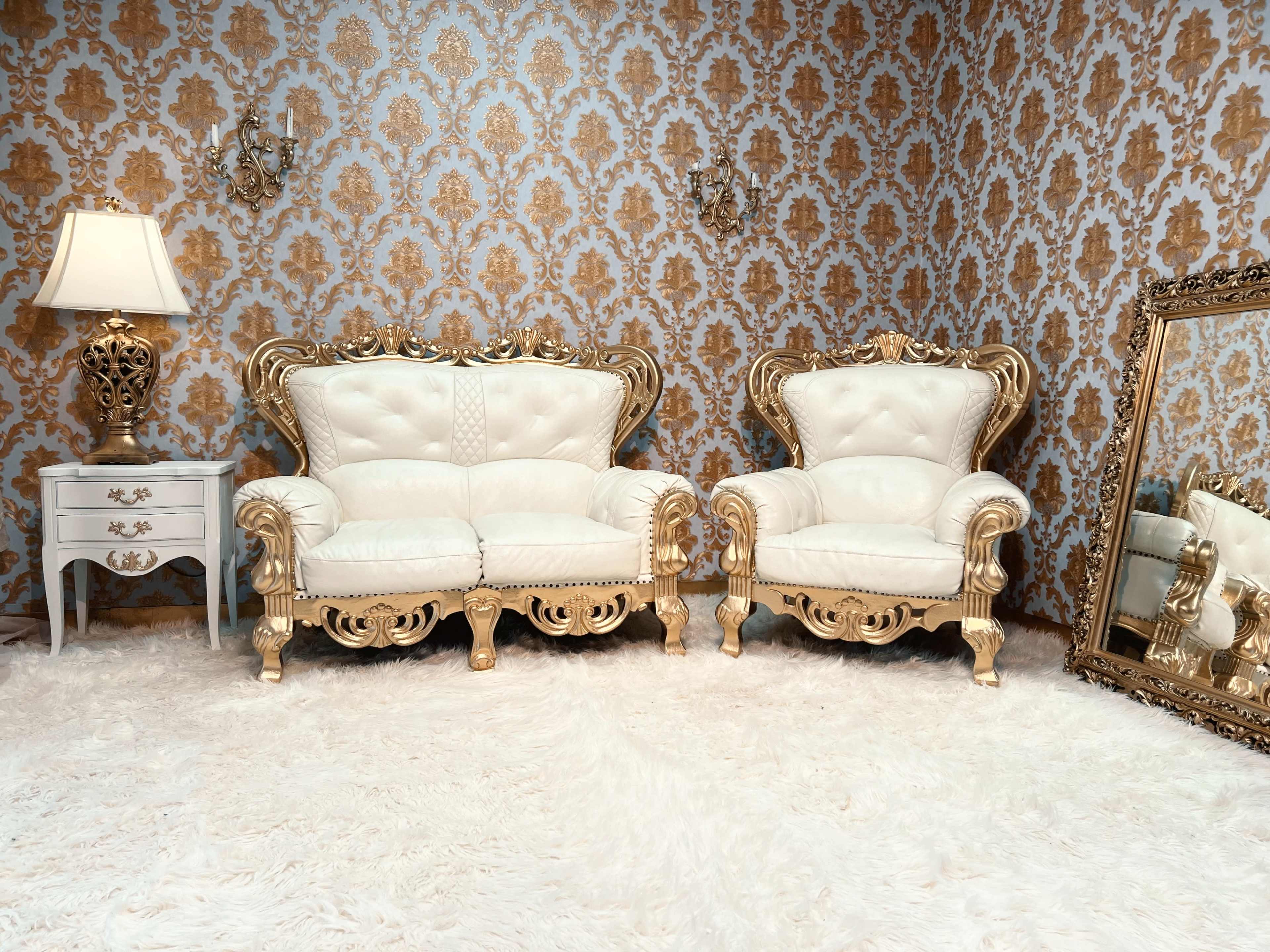 The image features two ornate white and gold upholstered chairs arranged opposite a decorative mirror, against a backdrop of golden damask wallpaper and a plush white rug.