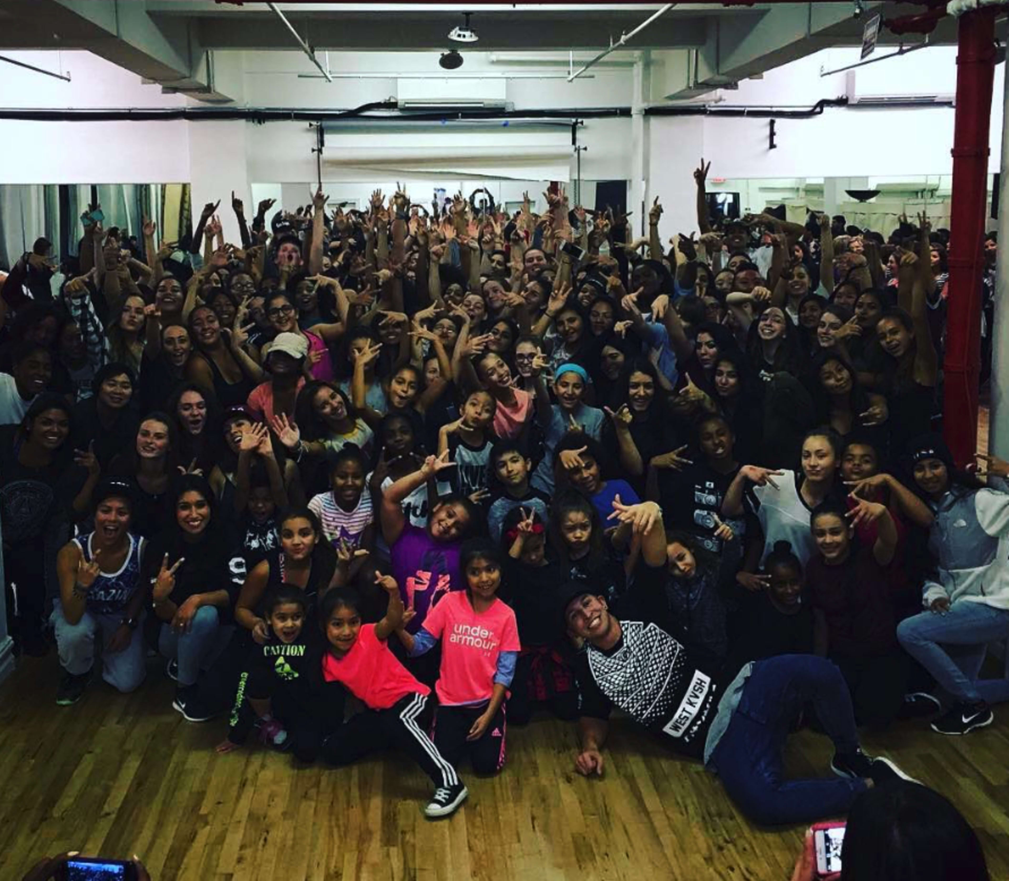 A large group of people, primarily young dancers, pose together with raised hands in a dance studio.