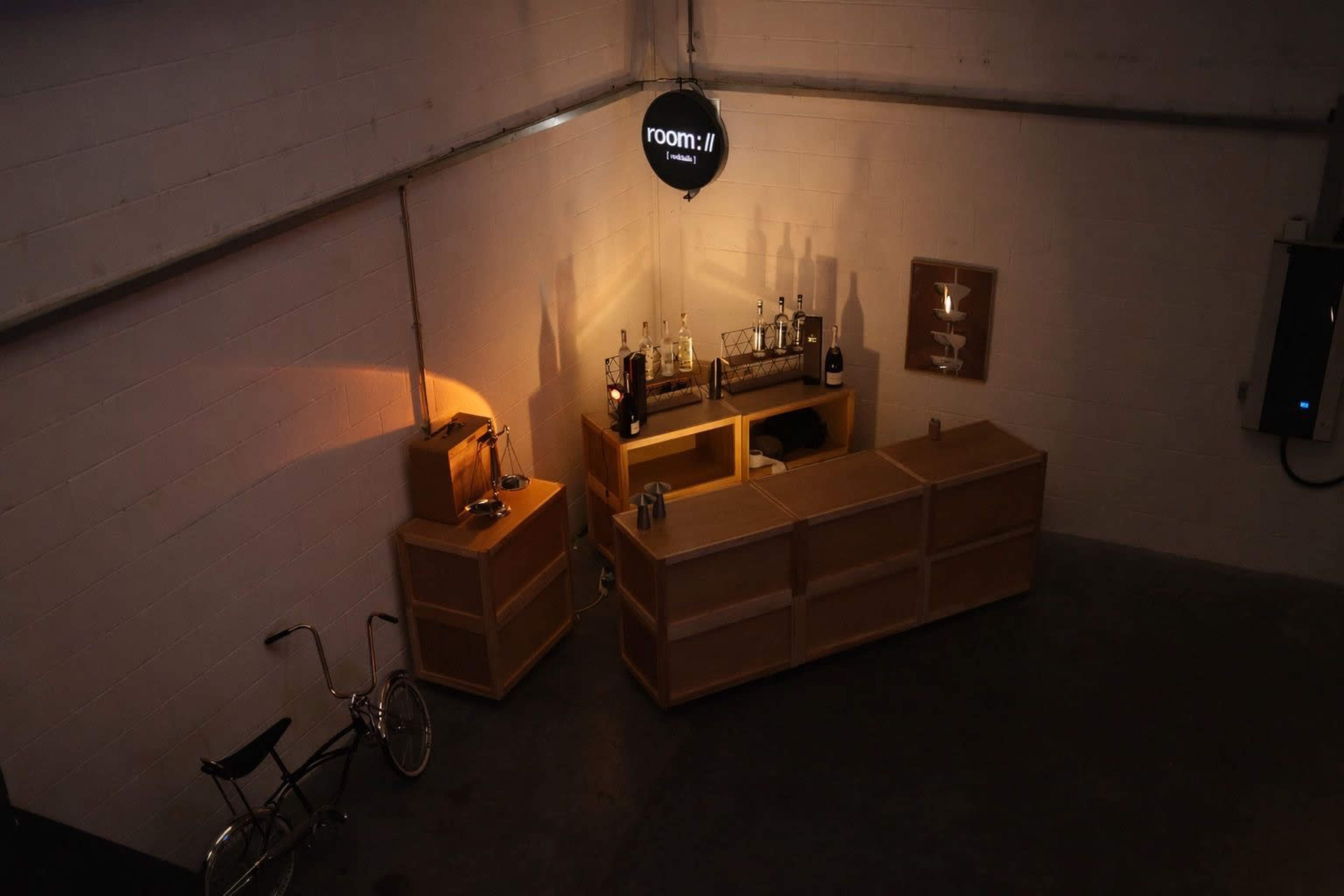 The image shows a minimalistic bar setup in a warehouse space, featuring wooden cabinets, bottles on display, and a bicycle in the corner.