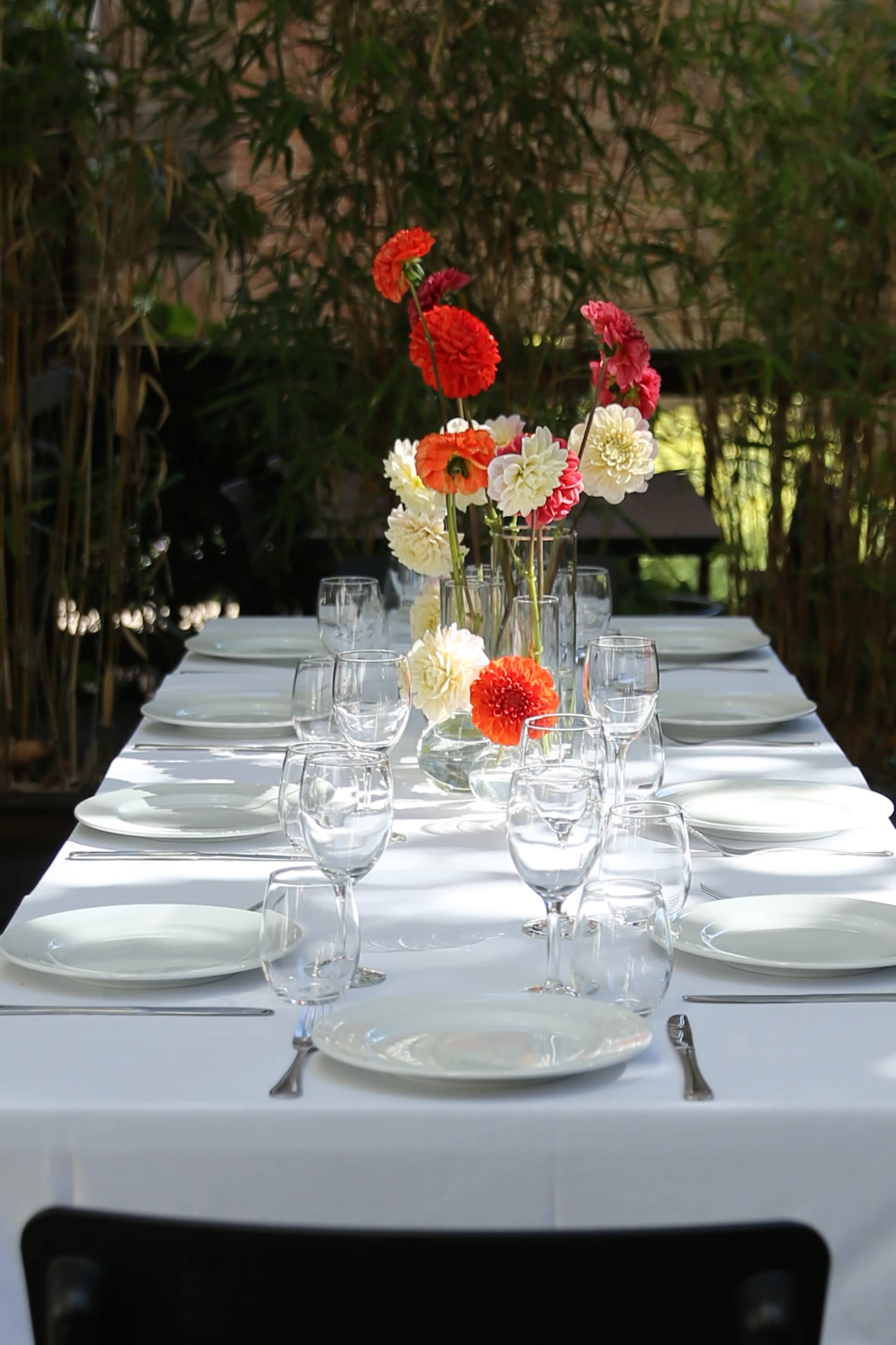 A long dining table is set with white plates and glassware, featuring a central arrangement of colorful flowers in vases.