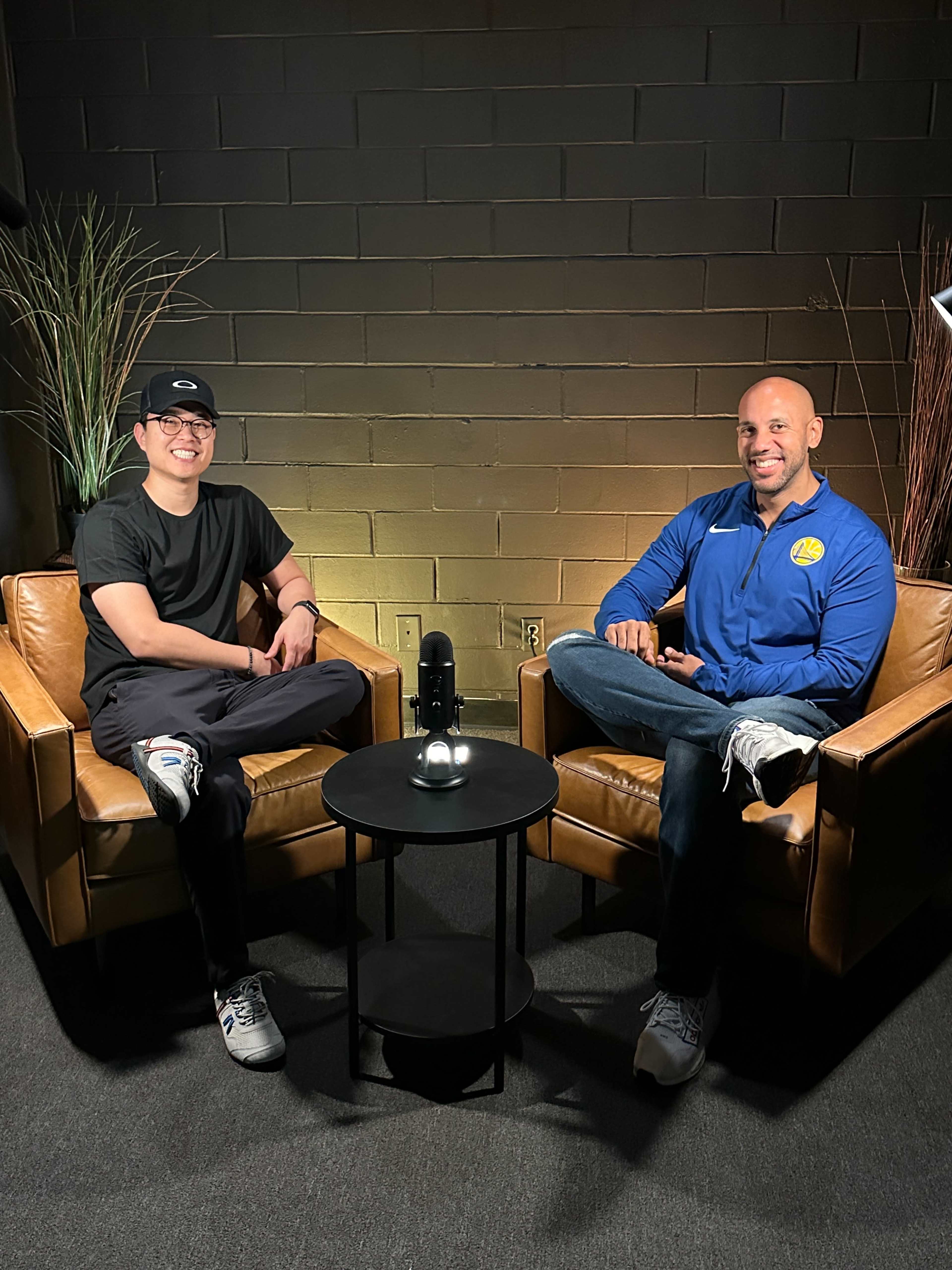 Two men sit in brown leather chairs facing each other in a dimly lit room, with a small round table and a microphone between them.
