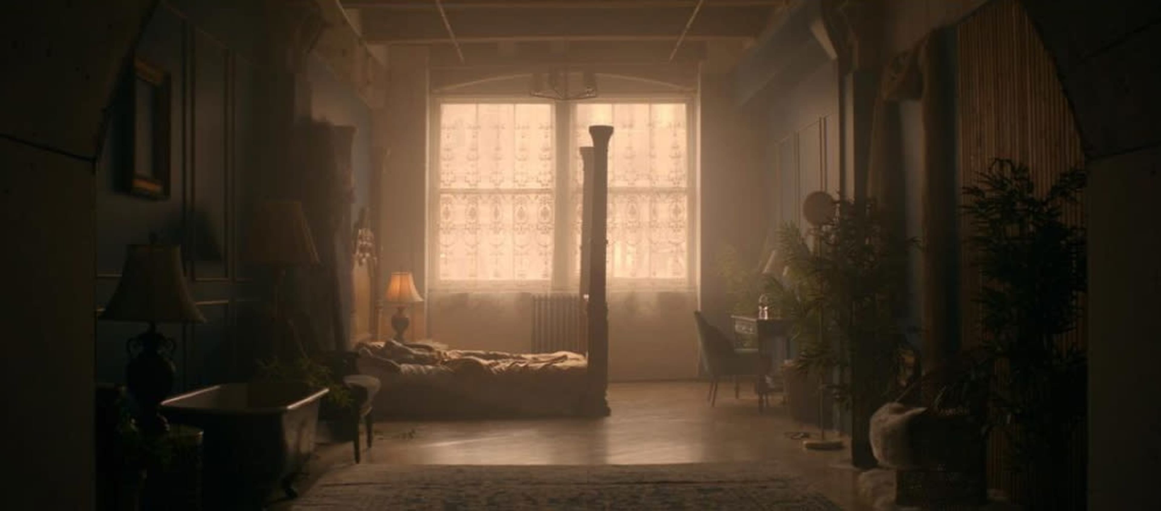 A softly lit room features a bed in the center, flanked by a window with sheer curtains and surrounded by plants and furniture.