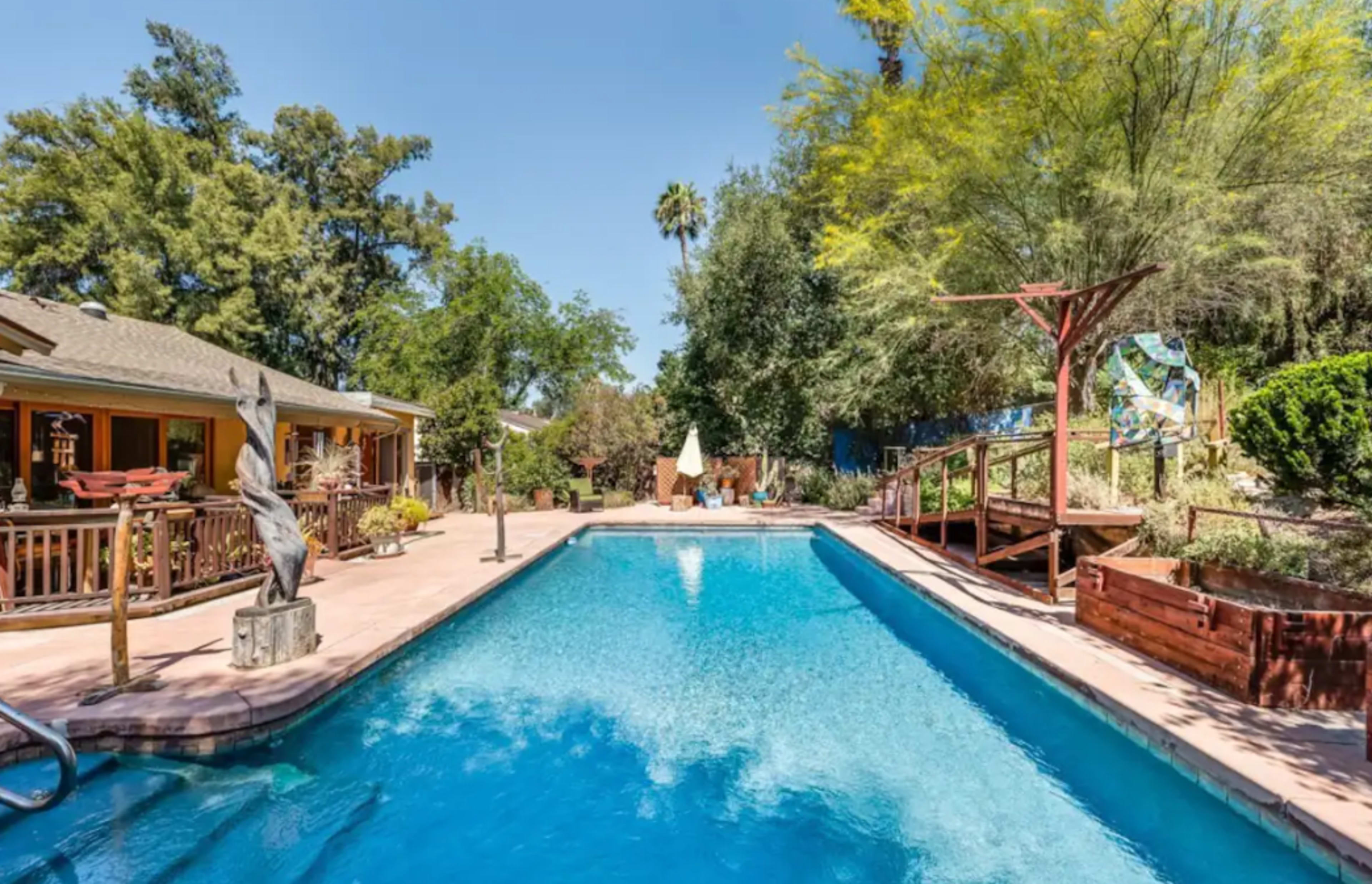 A rectangular swimming pool is surrounded by trees and landscaped areas, with a deck and seating areas visible in the background.
