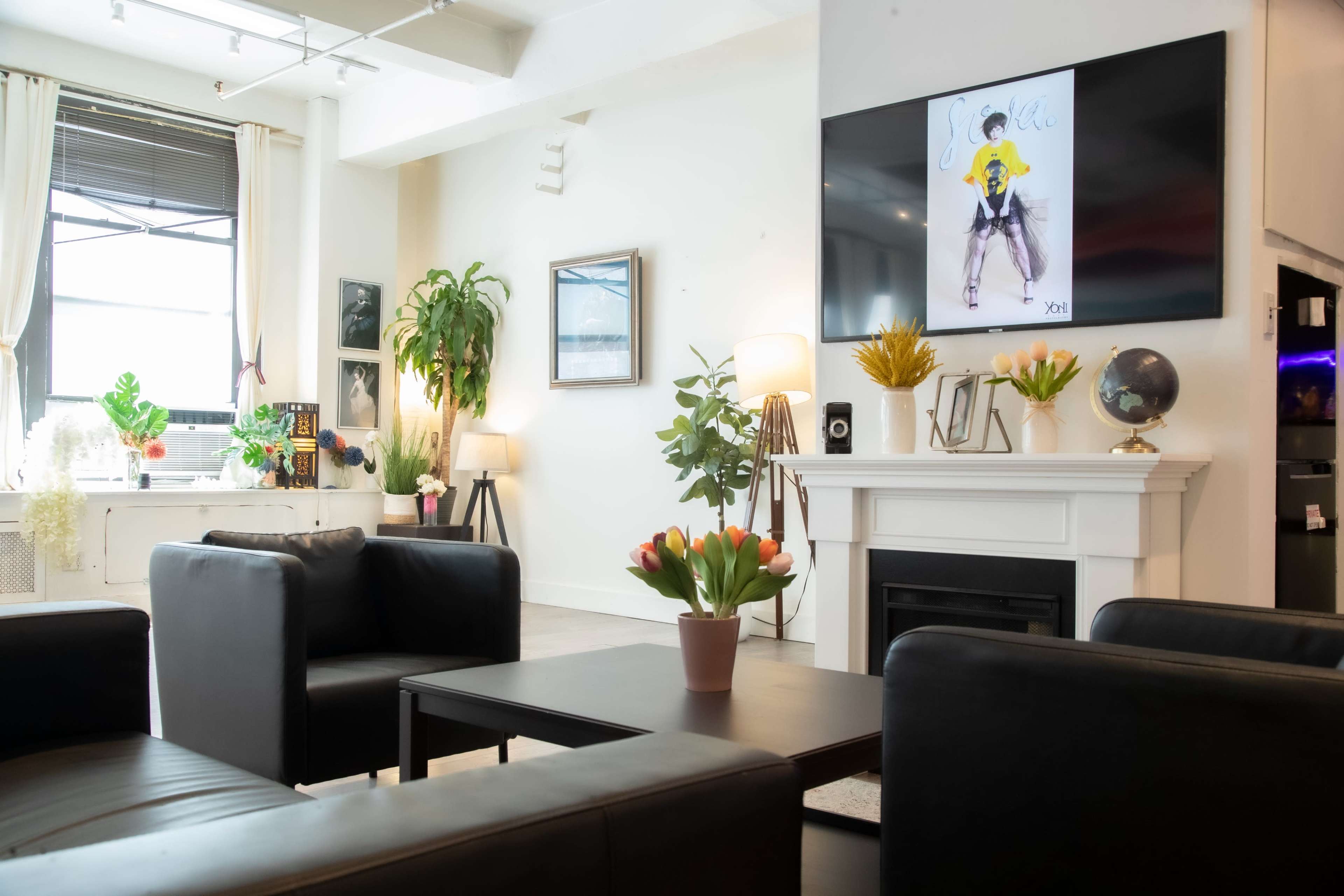 A modern living room with two black sofas facing a wooden coffee table, a fireplace with a large wall-mounted television above, and decorative plants and artwork on the walls.