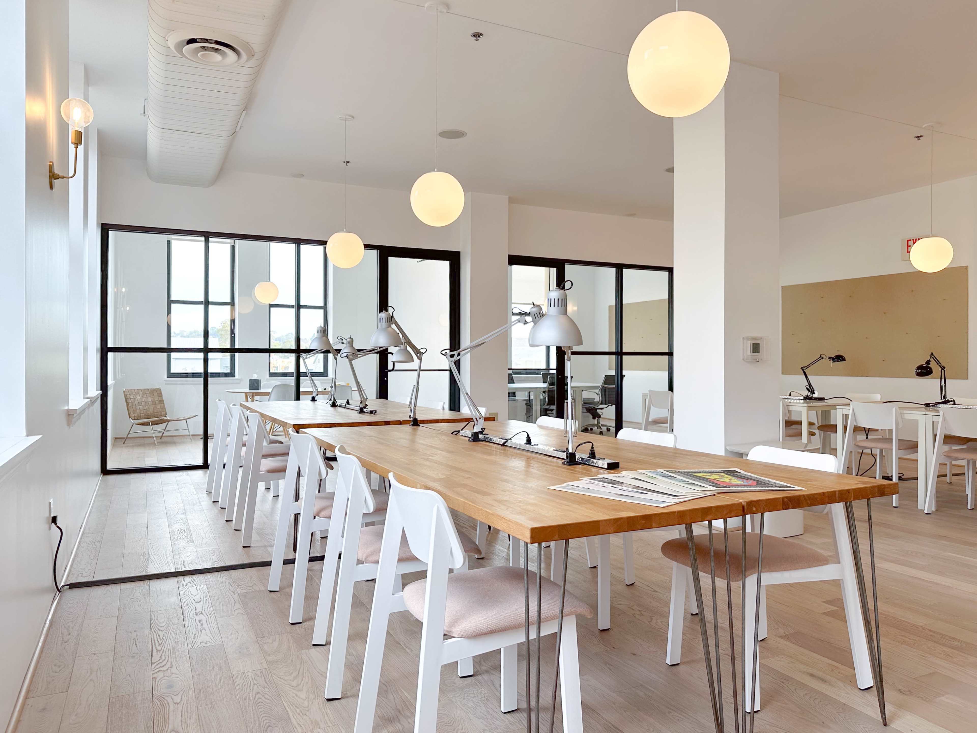 A modern, well-lit workspace featuring a long wooden table surrounded by white chairs and several task lamps, with large windows allowing natural light to enter.