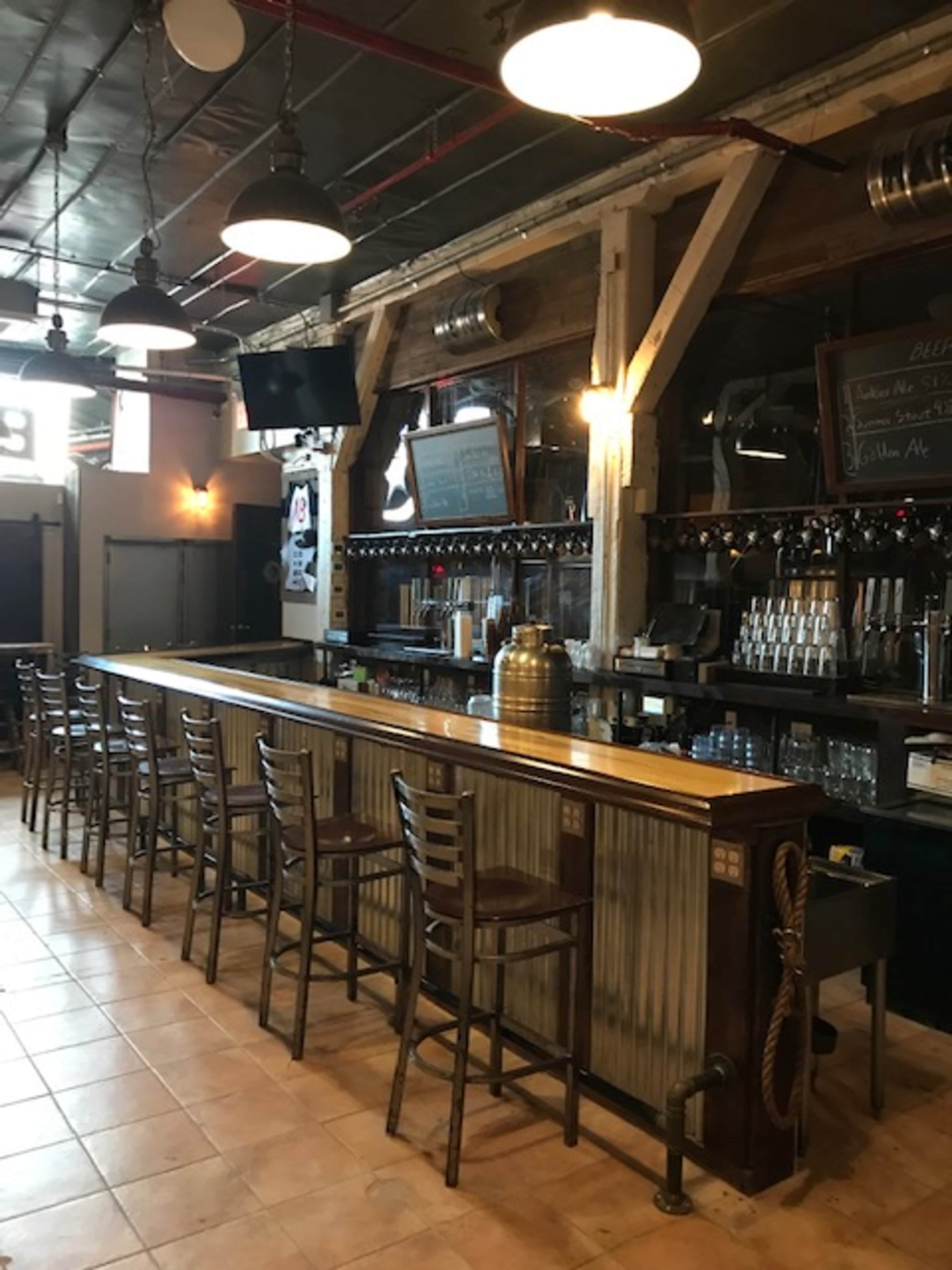 A wooden bar with metal accents and high stools, illuminated by overhead lights in a rustic setting.