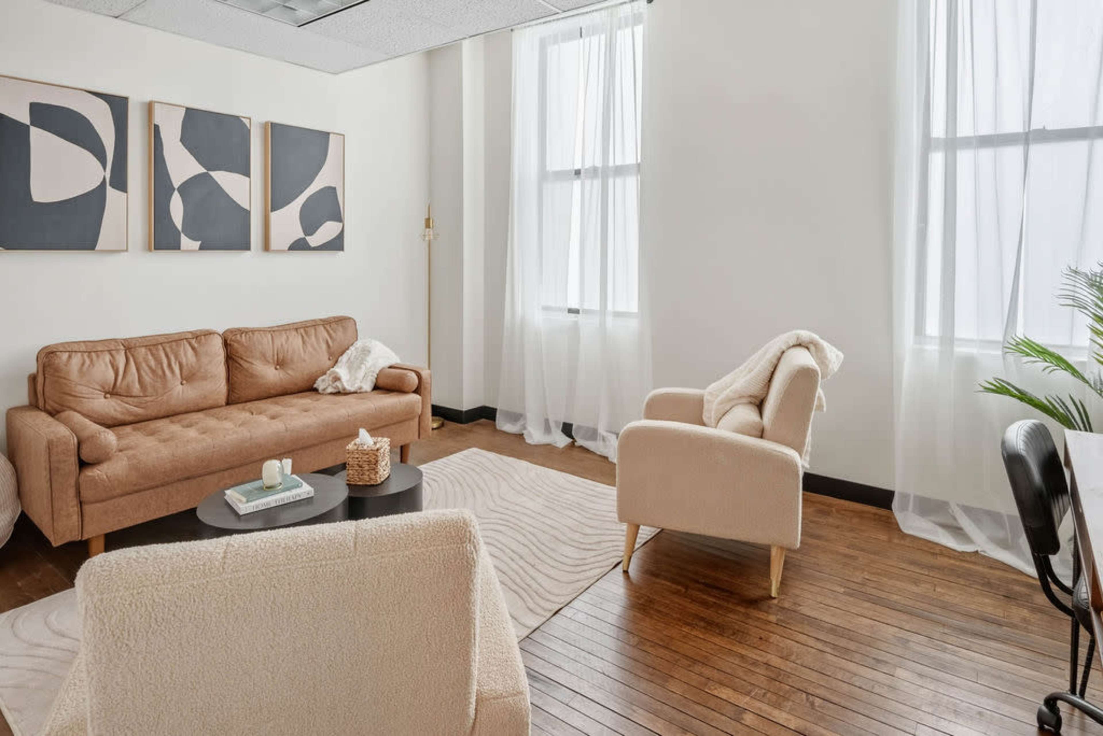 The image shows a modern, minimalistic living space with a brown leather sofa, two light-colored armchairs, and abstract artwork on the walls, illuminated by natural light from large windows.