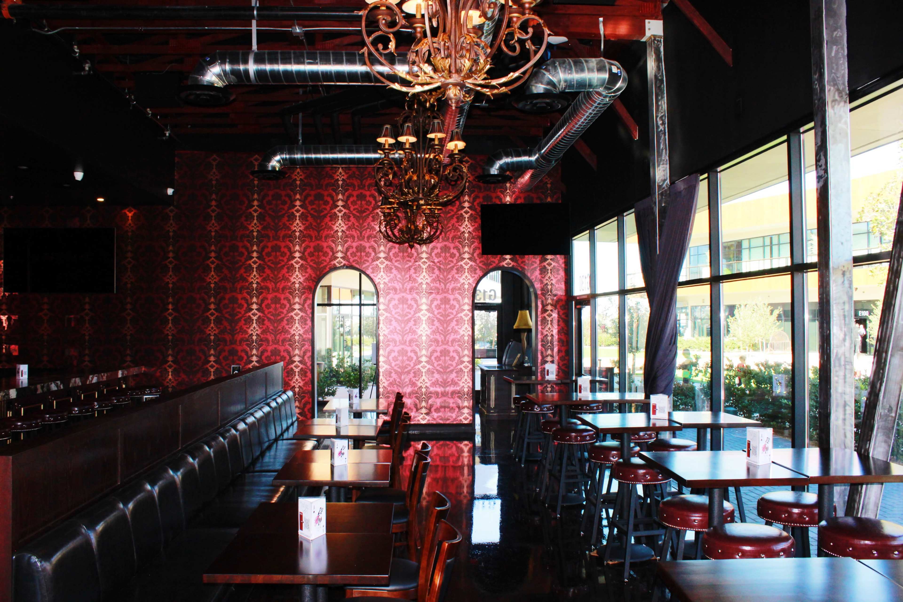 The image shows an interior of a restaurant featuring dark wooden tables and chairs, a patterned wallpaper, and large windows letting in natural light.