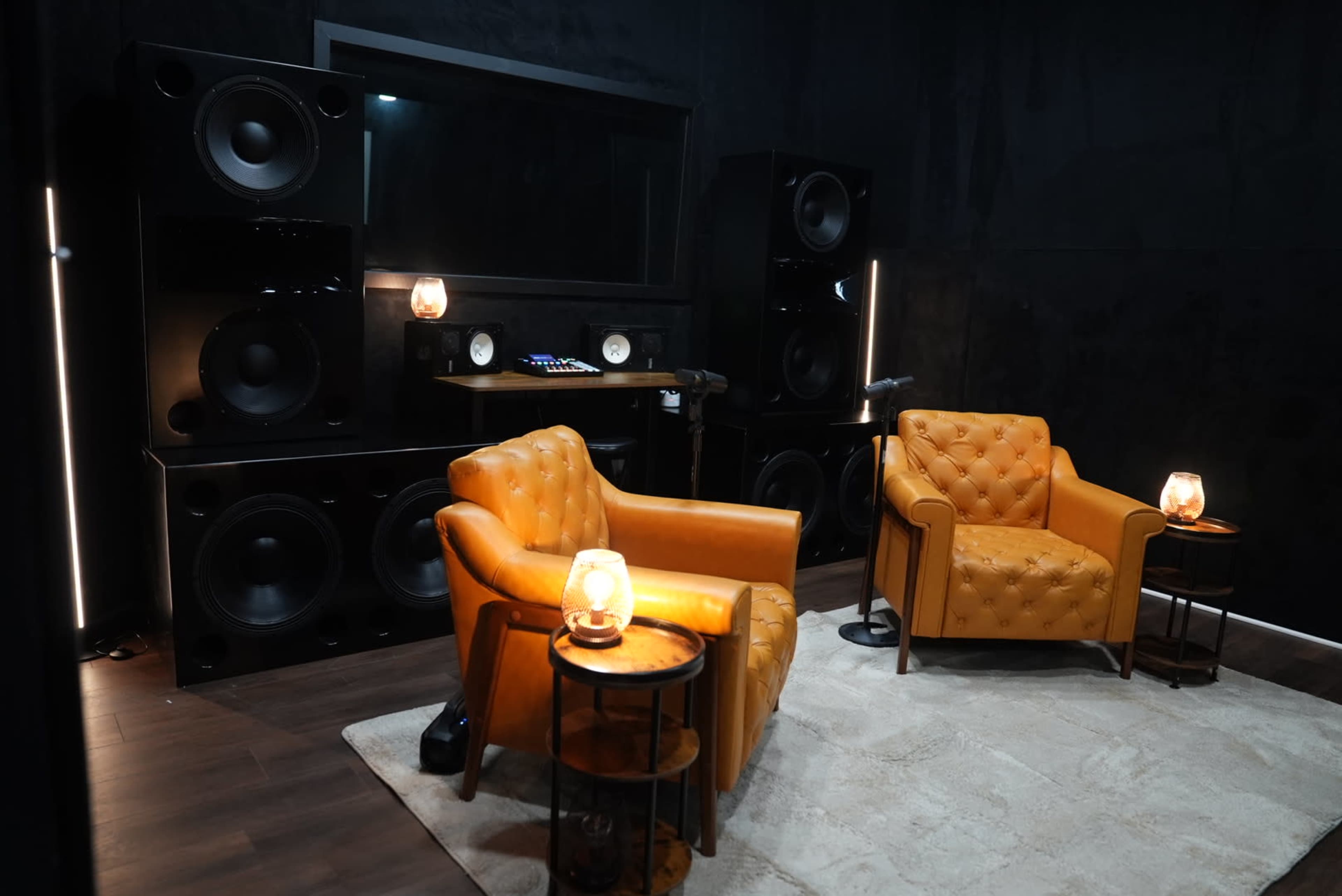 The image shows a sound studio featuring two orange upholstered chairs, a pair of large black speakers, and a desk with audio equipment, illuminated by soft lighting.