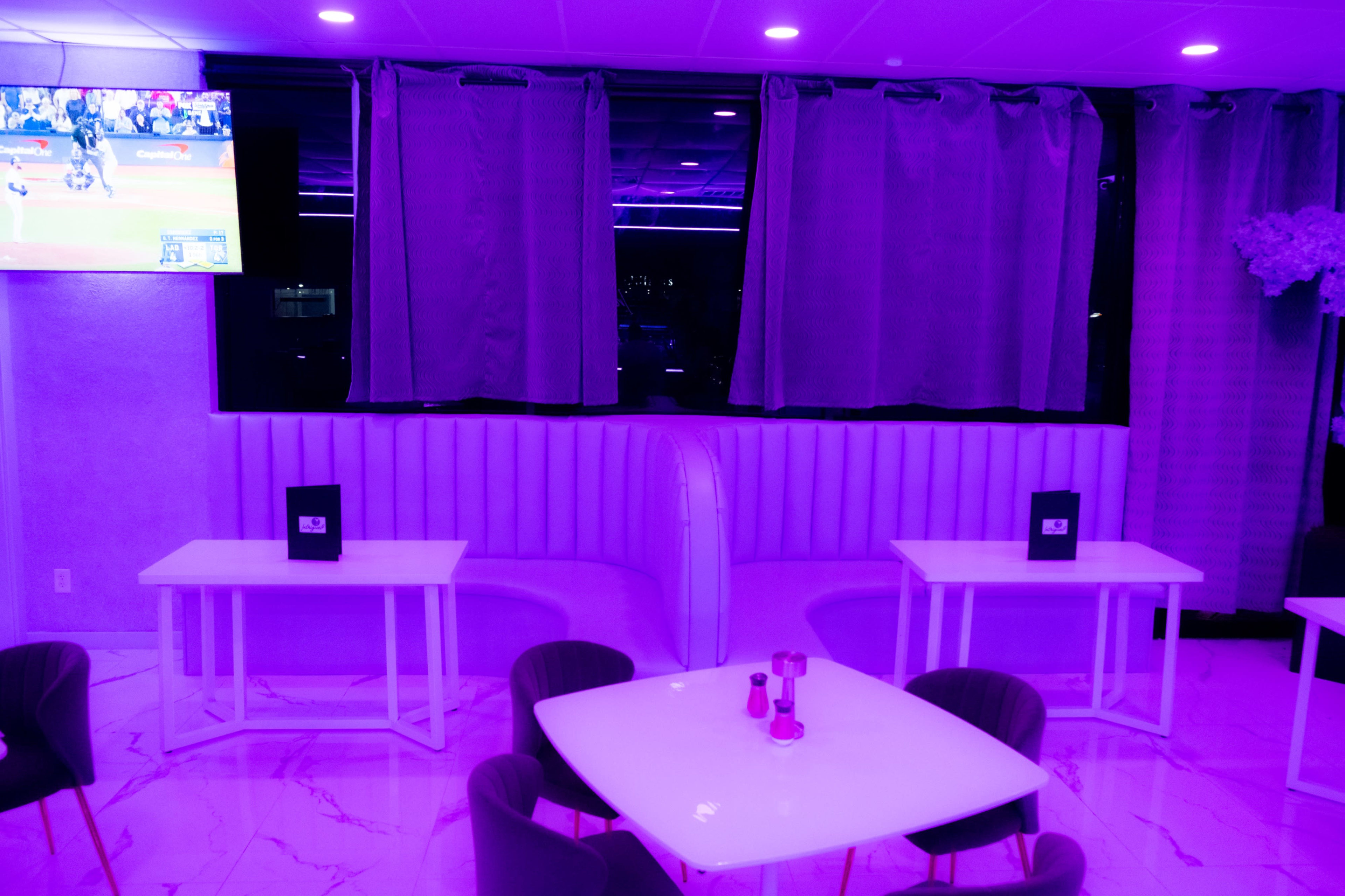 A restaurant interior features white tables and chairs under purple lighting, with a large TV displaying a sports game in the background and partially drawn curtains.