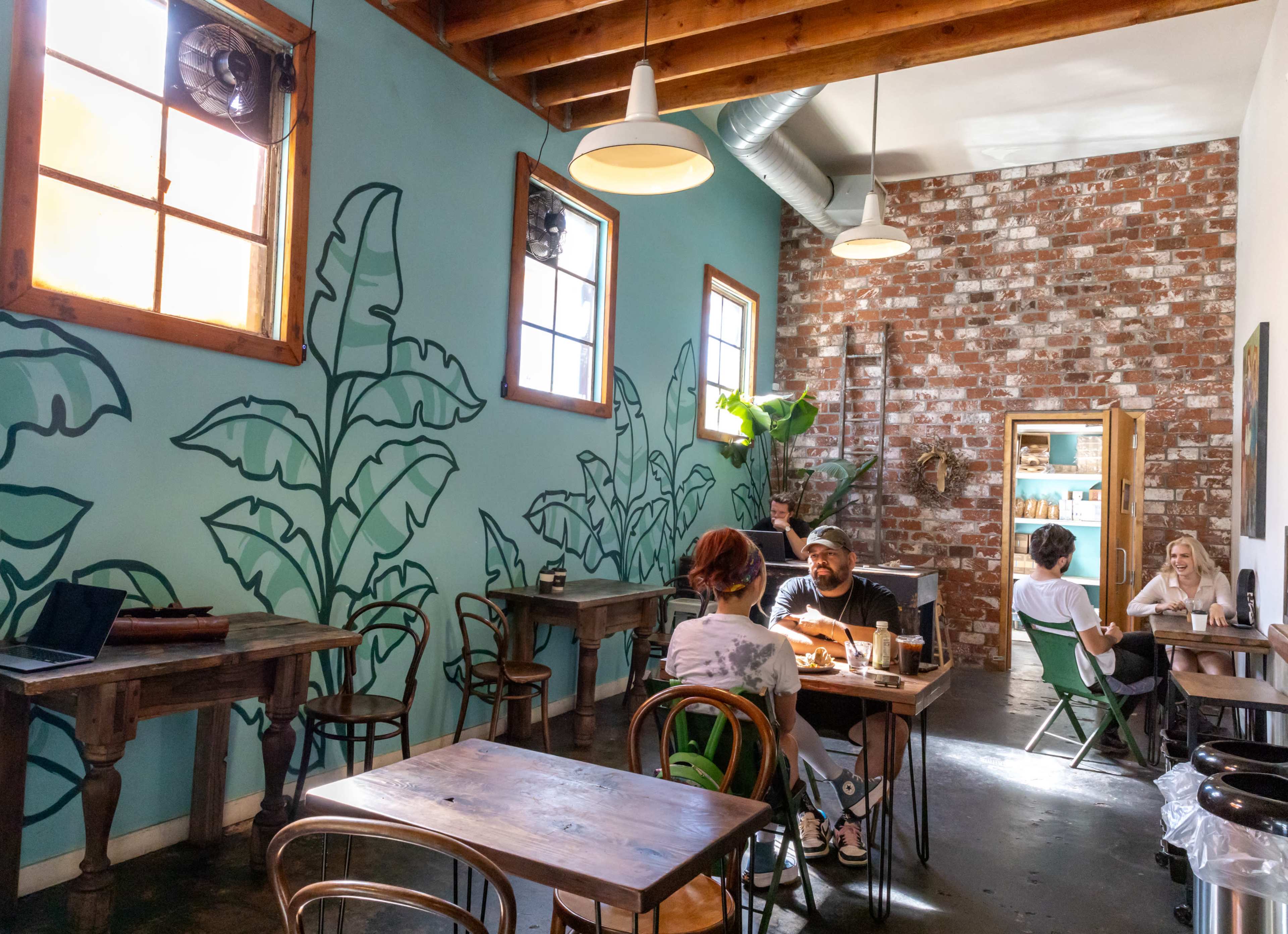A cozy café interior with a brick wall, wooden tables, and walls painted with large green plant illustrations.