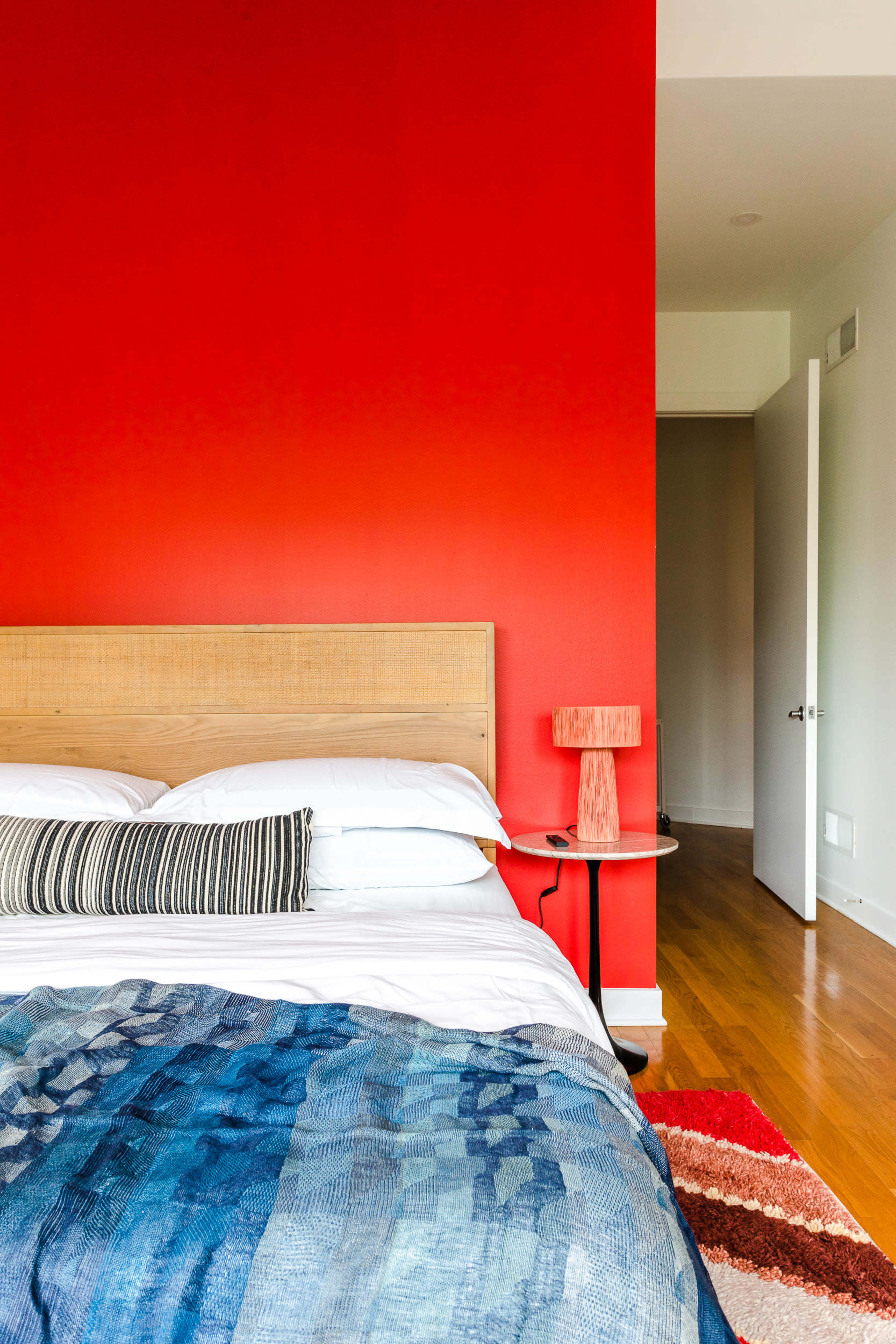 A bed with a patterned blanket and a striped pillow is positioned against a bright red wall, next to a wooden bedside table and a doorway leading to another room.