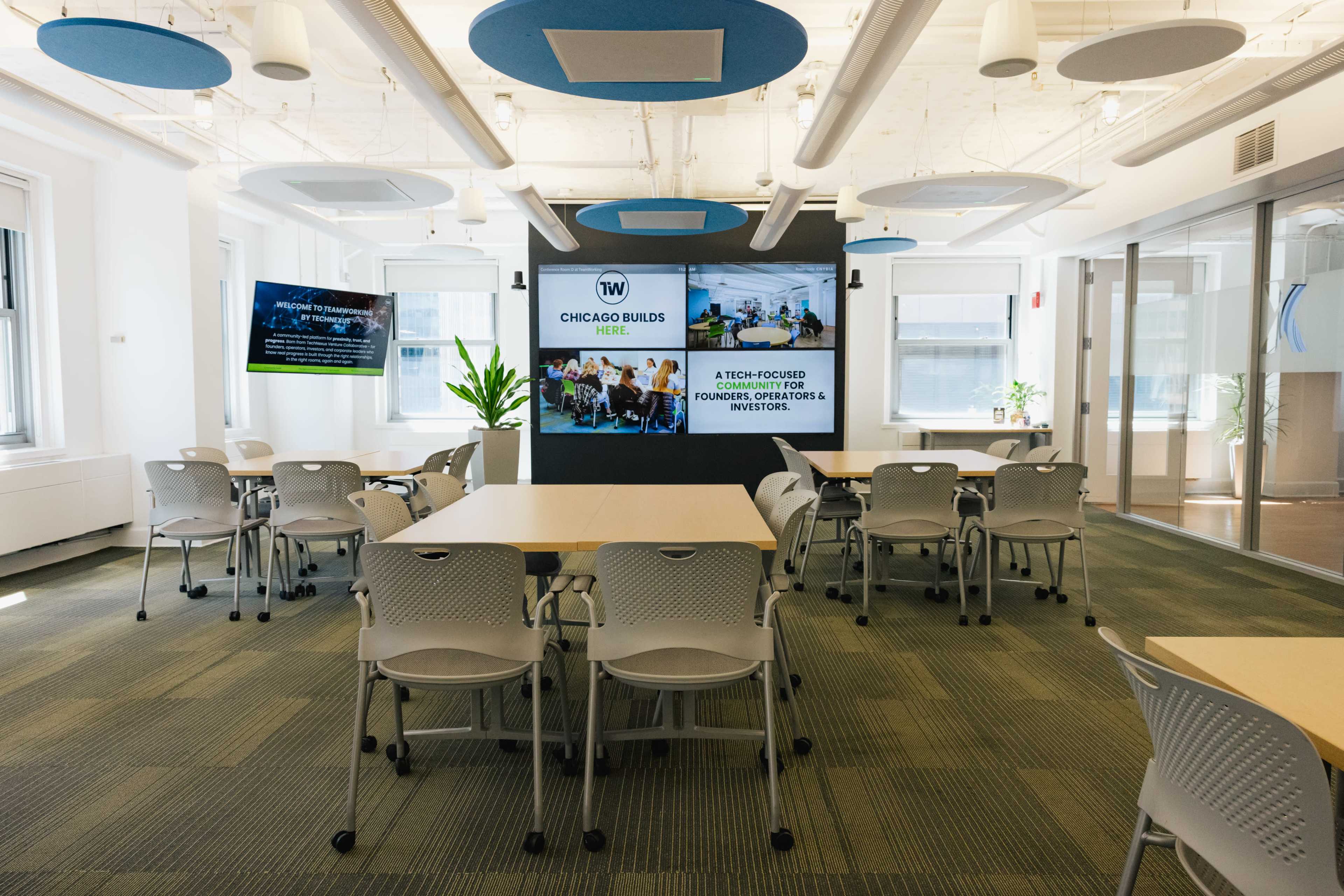 The image shows a modern office space with several tables and chairs arranged for collaboration, featuring large screens displaying information about a program called "Chicago Builds."