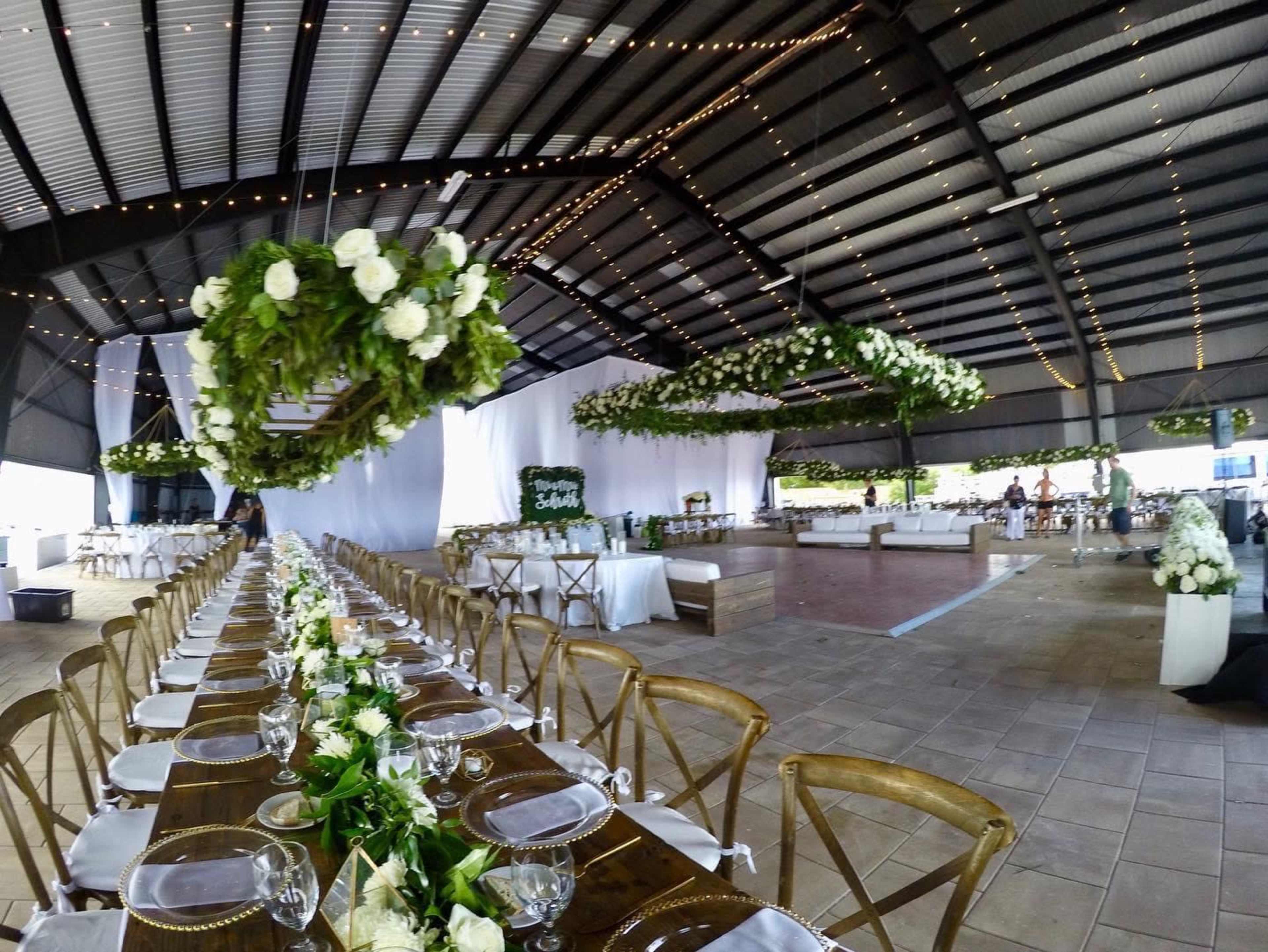 The image shows a large indoor event space with long dining tables elegantly arranged and decorated with white flowers and greenery, under a canopy of string lights.