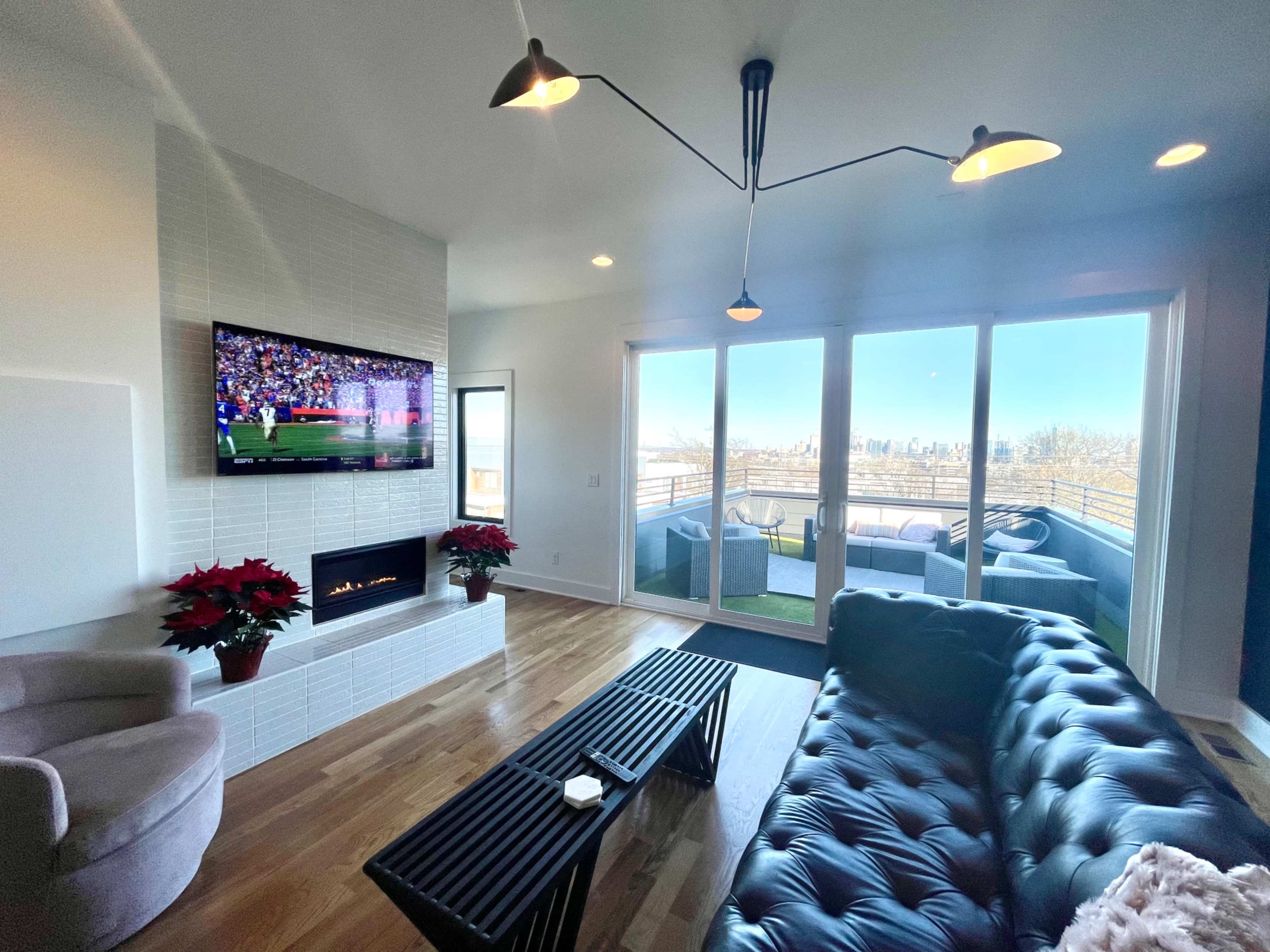 A modern living room features a wall-mounted TV, a fireplace, a dark sofa, and large windows that offer a view of a city skyline.