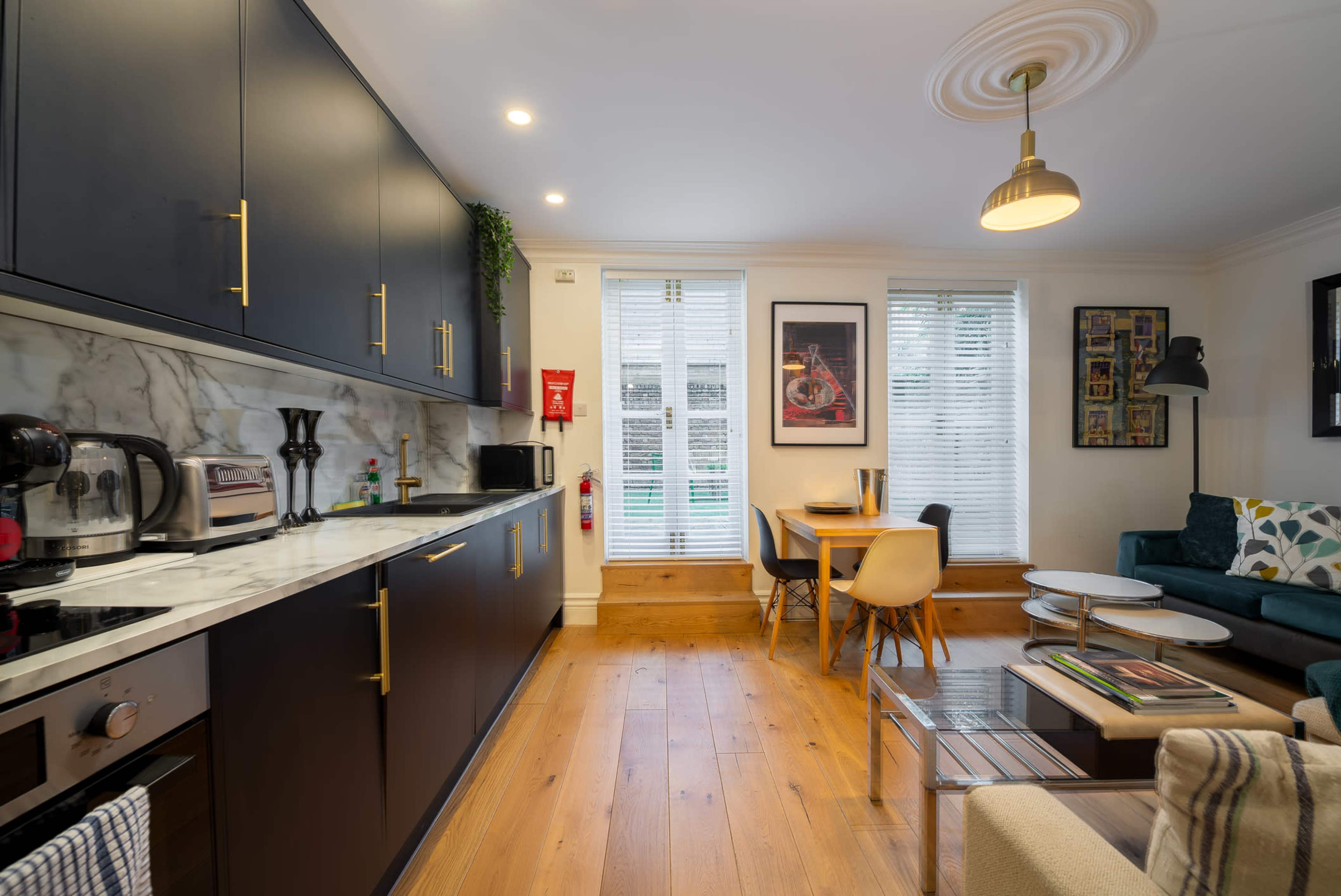 The image shows a modern kitchen and living area featuring dark cabinetry, a marble countertop, and a small dining table beside large windows with white blinds.