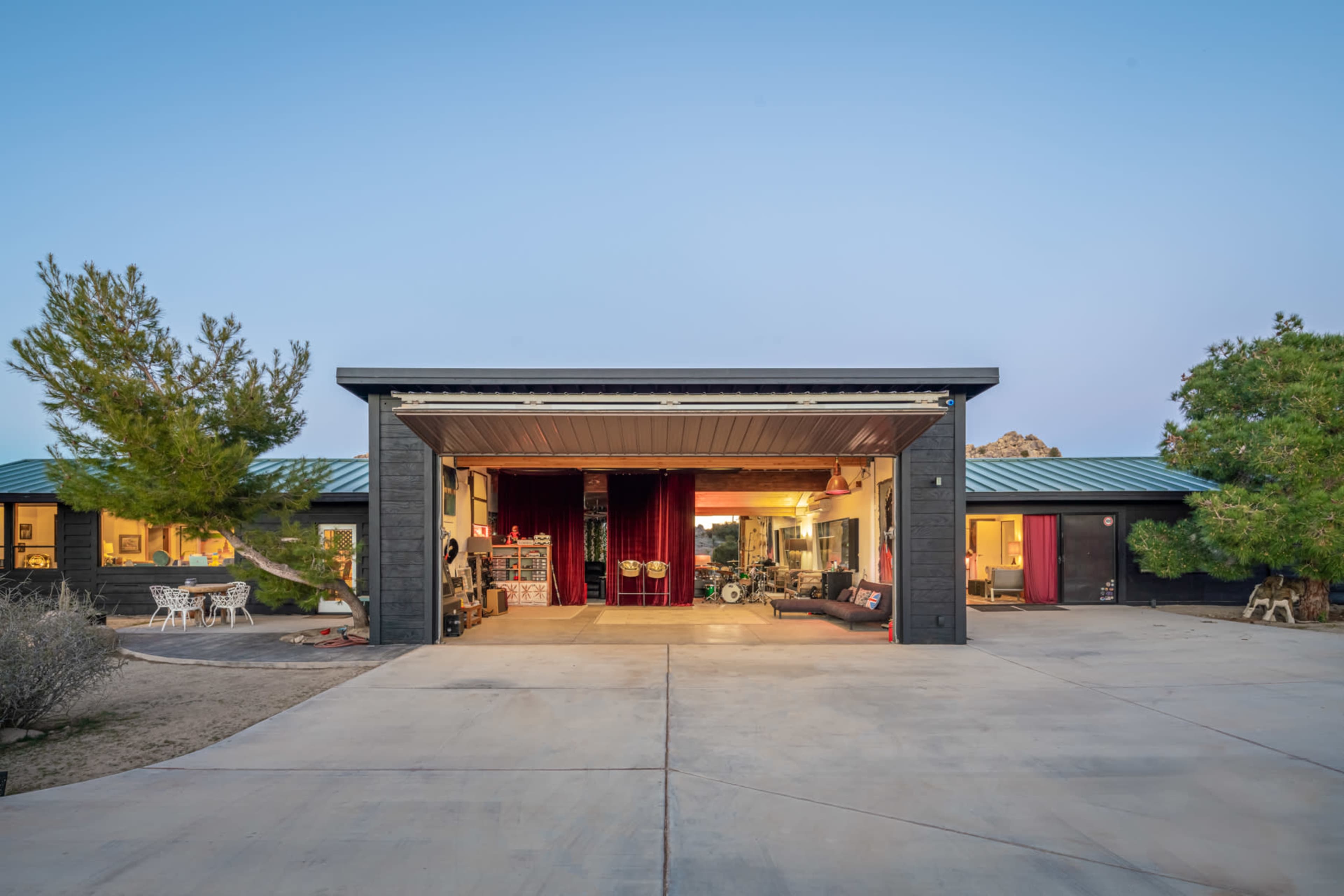 A modern, open-concept home with a large roll-up garage door facing a paved driveway and surrounded by desert vegetation.
