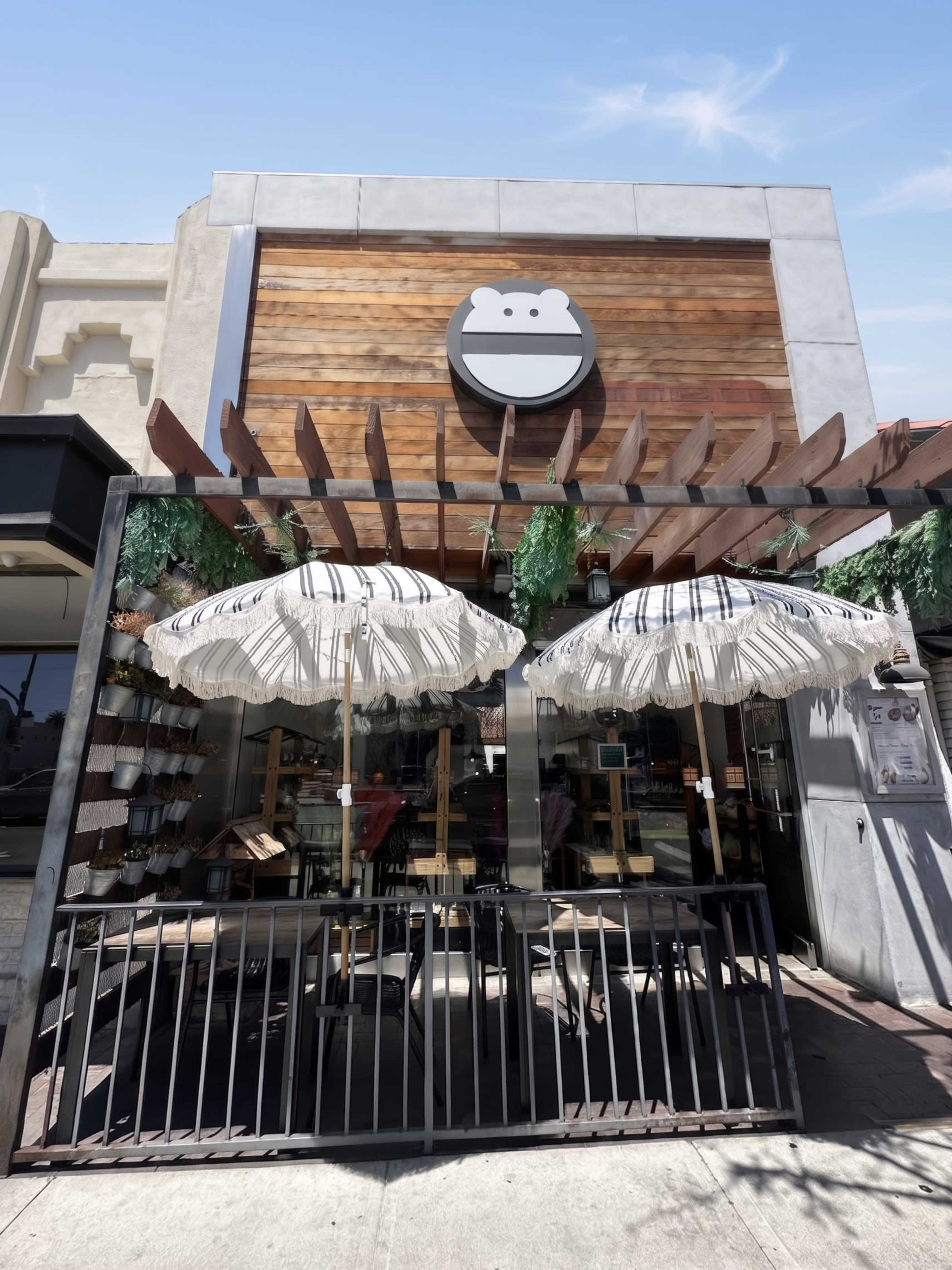 The image shows a restaurant exterior featuring a wooden facade, a large circular sign, and two striped umbrellas over a patio area with a fence.