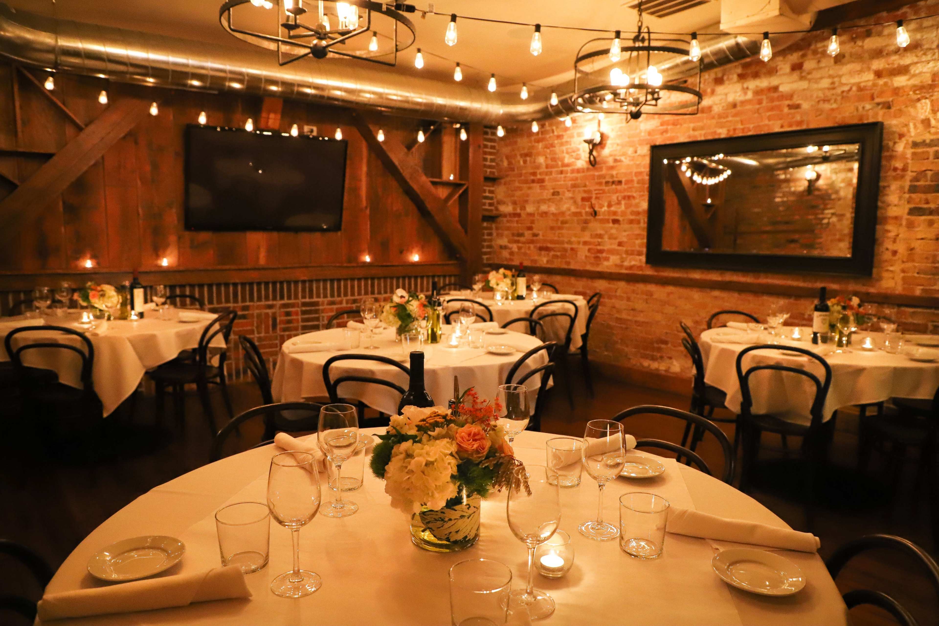 The image shows a cozy restaurant interior with wooden walls, round tables set for dining, and warm lighting from hanging bulbs.