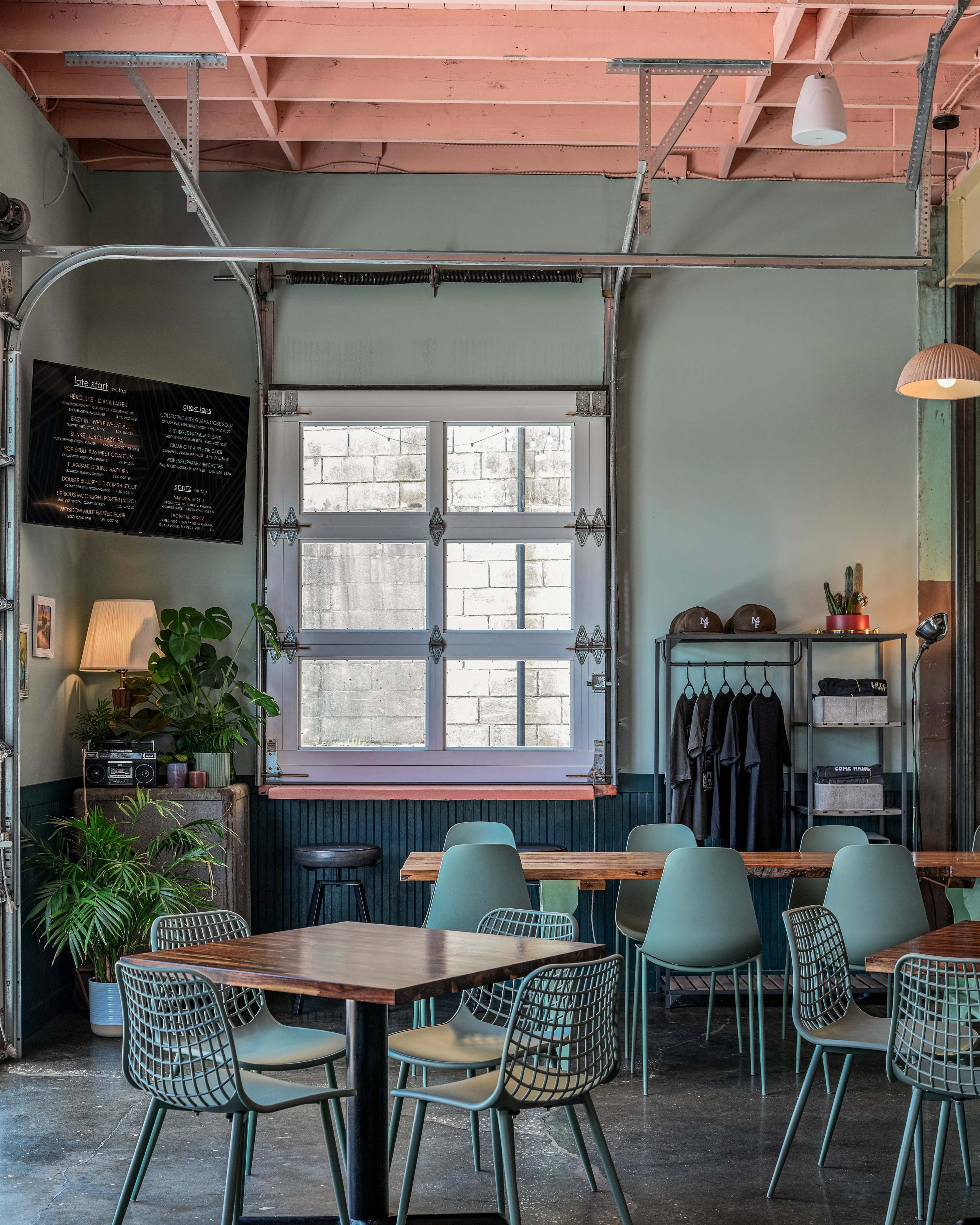 The image shows a modern café interior with green chairs, wooden tables, a large window, and plants.