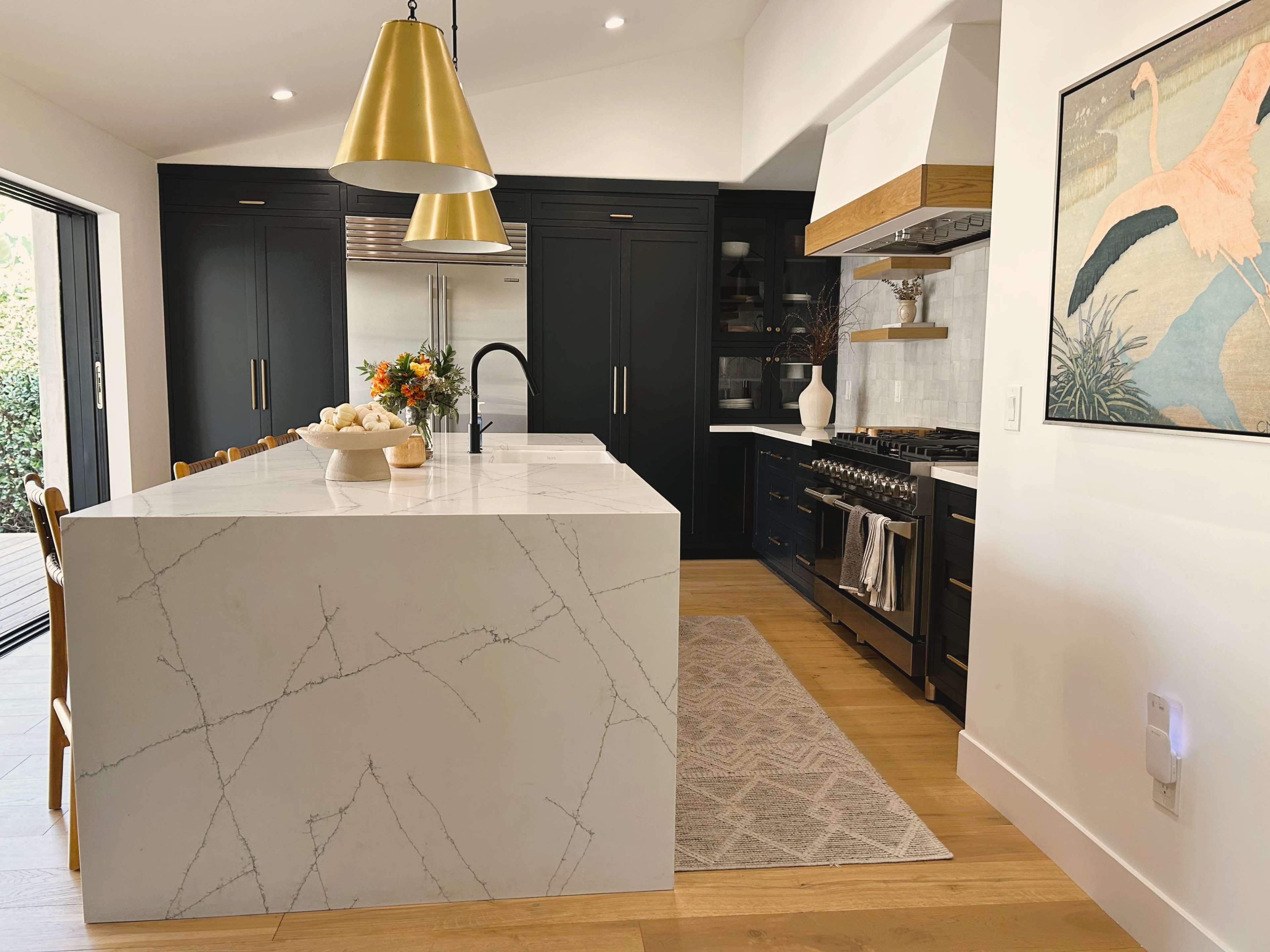 A modern kitchen features a large marble island with a bowl of fruit, black cabinetry, and stainless steel appliances.