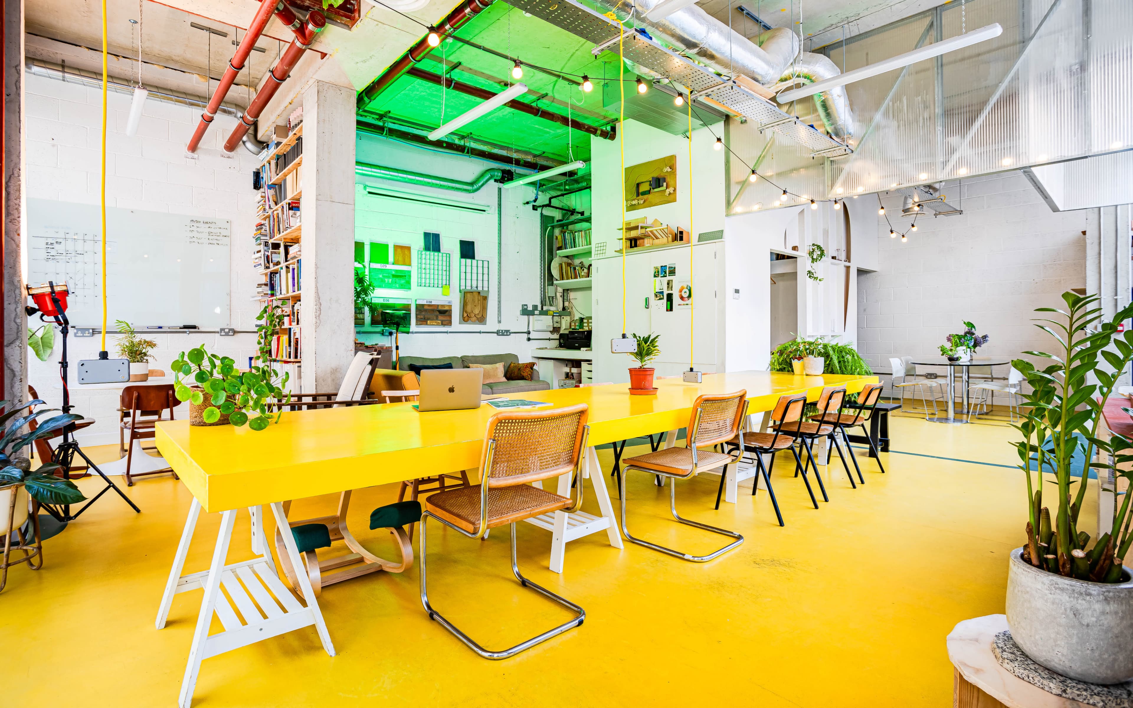 The image shows a bright, modern coworking space featuring a large yellow table surrounded by various chairs, lush plants, and an open layout with exposed industrial elements.