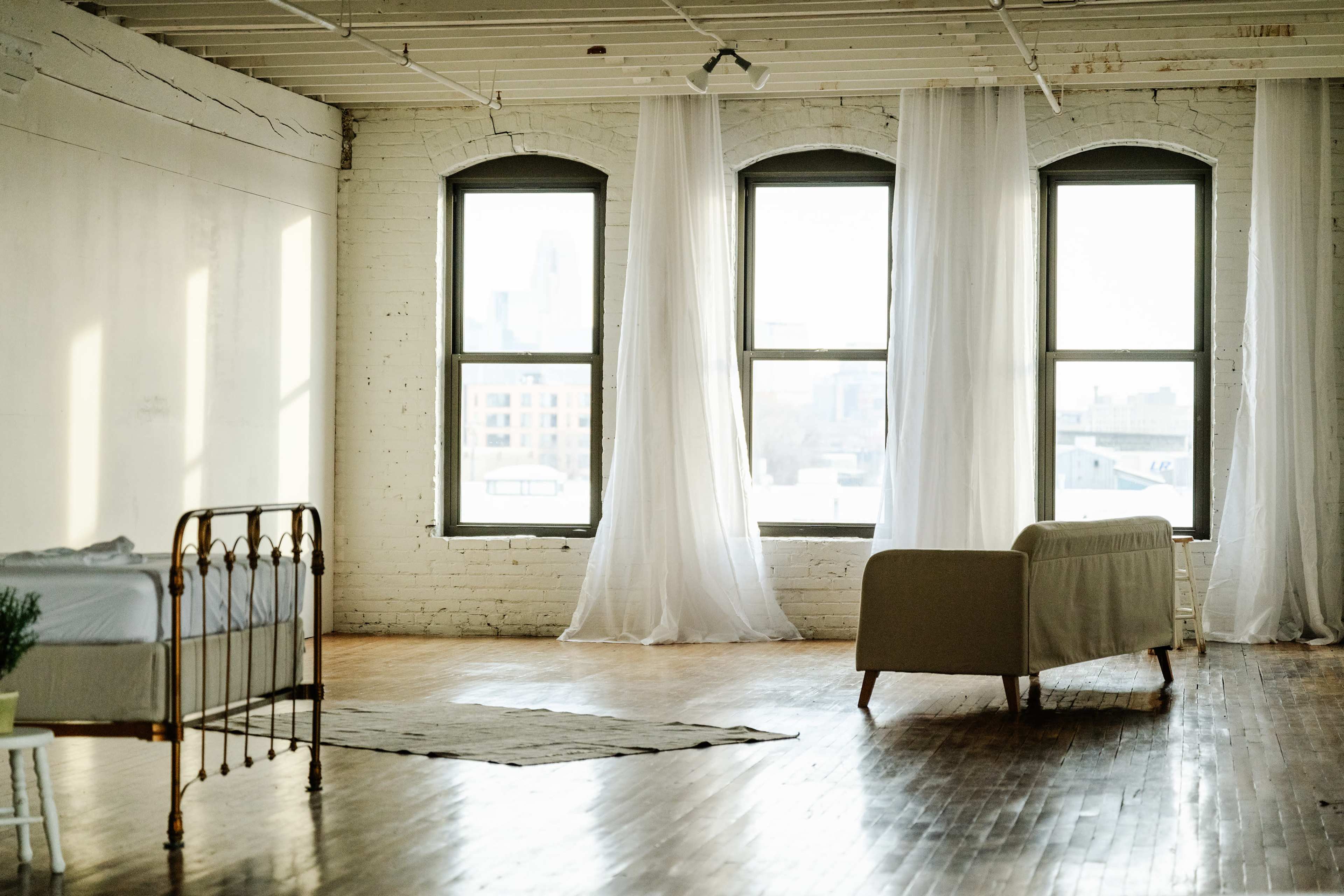 A spacious room with wooden flooring, two large windows framed by sheer white curtains, a bed with a metal frame, and a couch arranged beside a decorative rug.