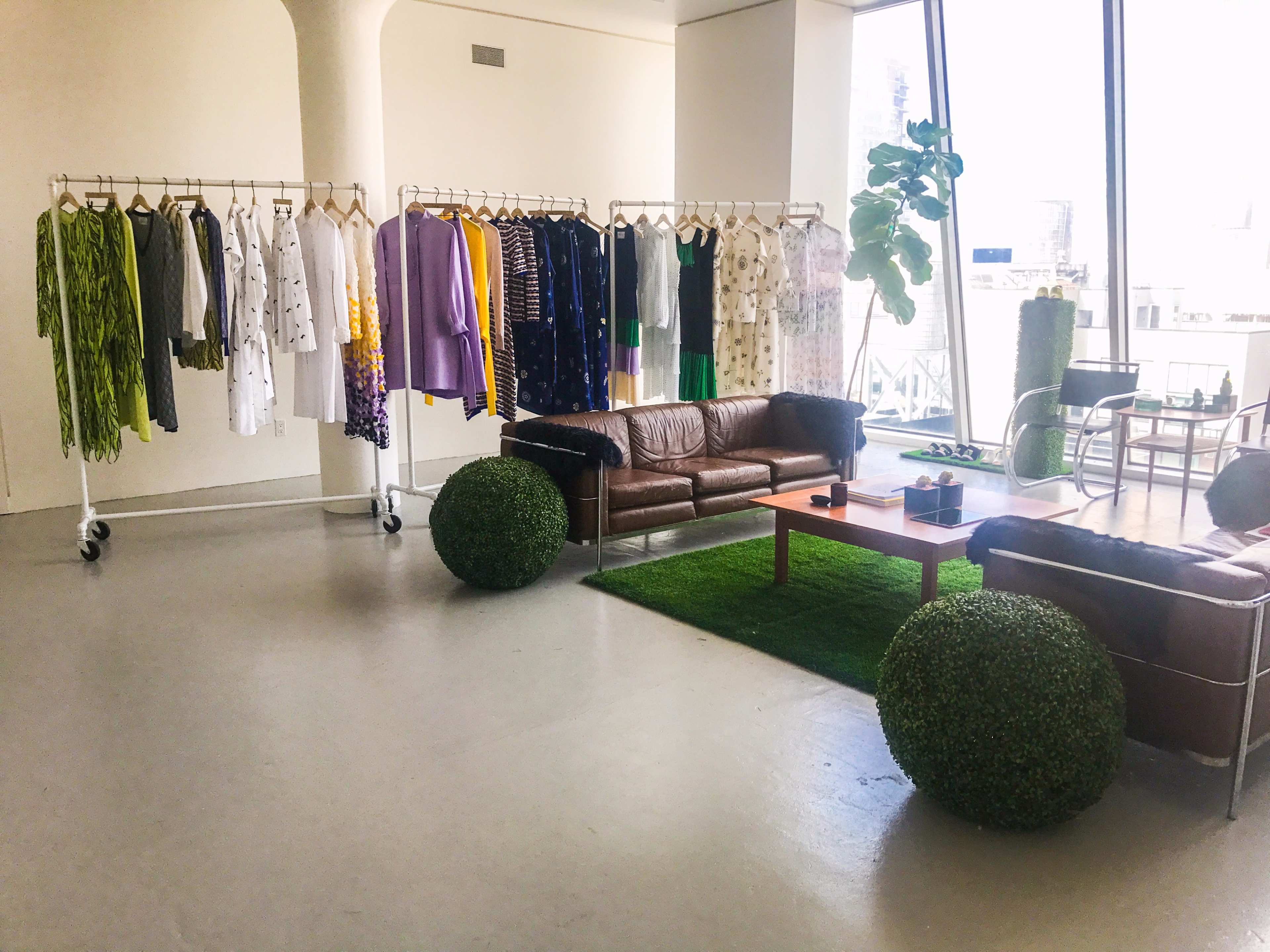 A bright room with racks of clothing in various colors, two brown leather sofas, a green area rug, and large windows.