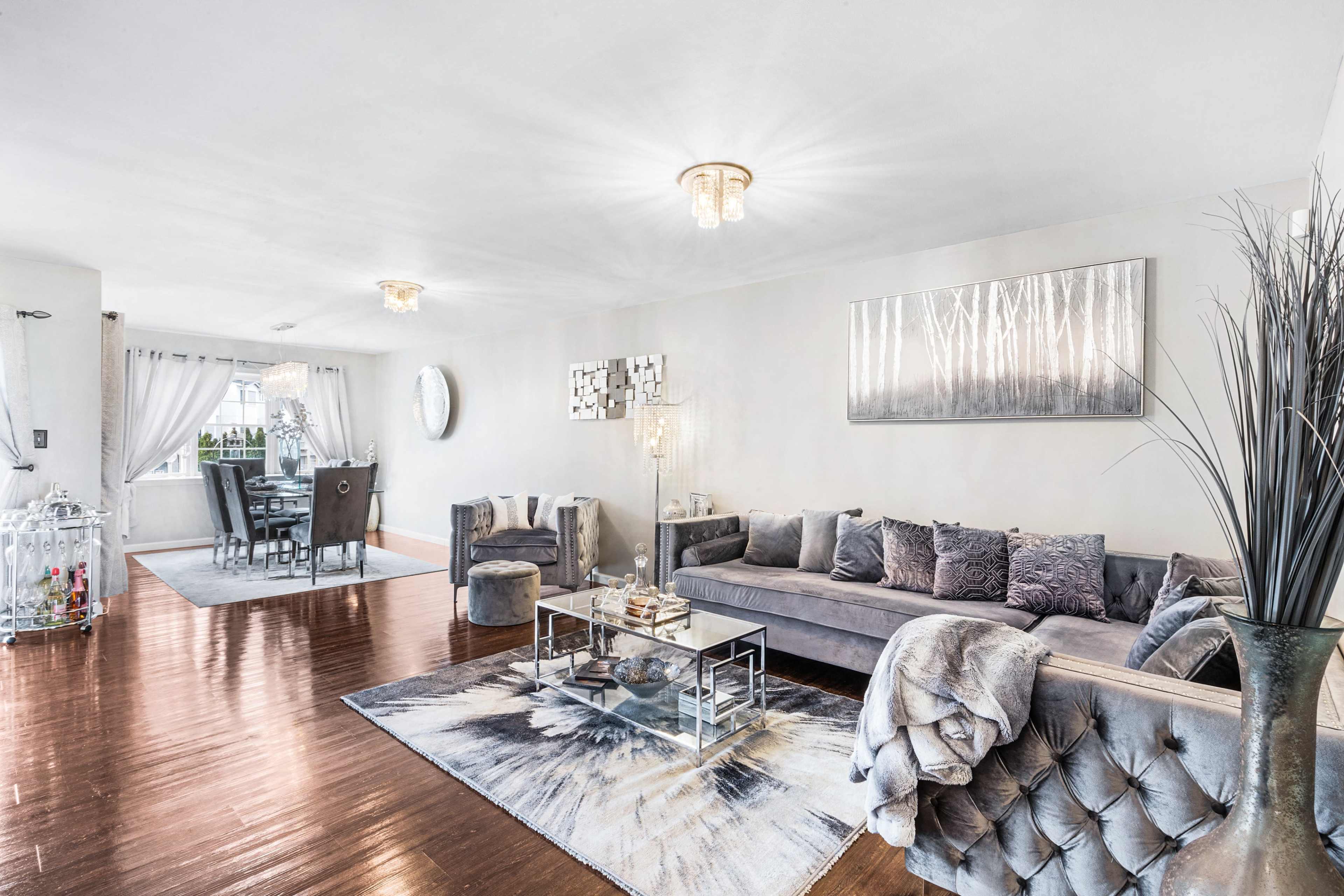 The image shows a modern living room with a gray sectional sofa, glass coffee table, and decorative artwork, opening to a dining area in the background.