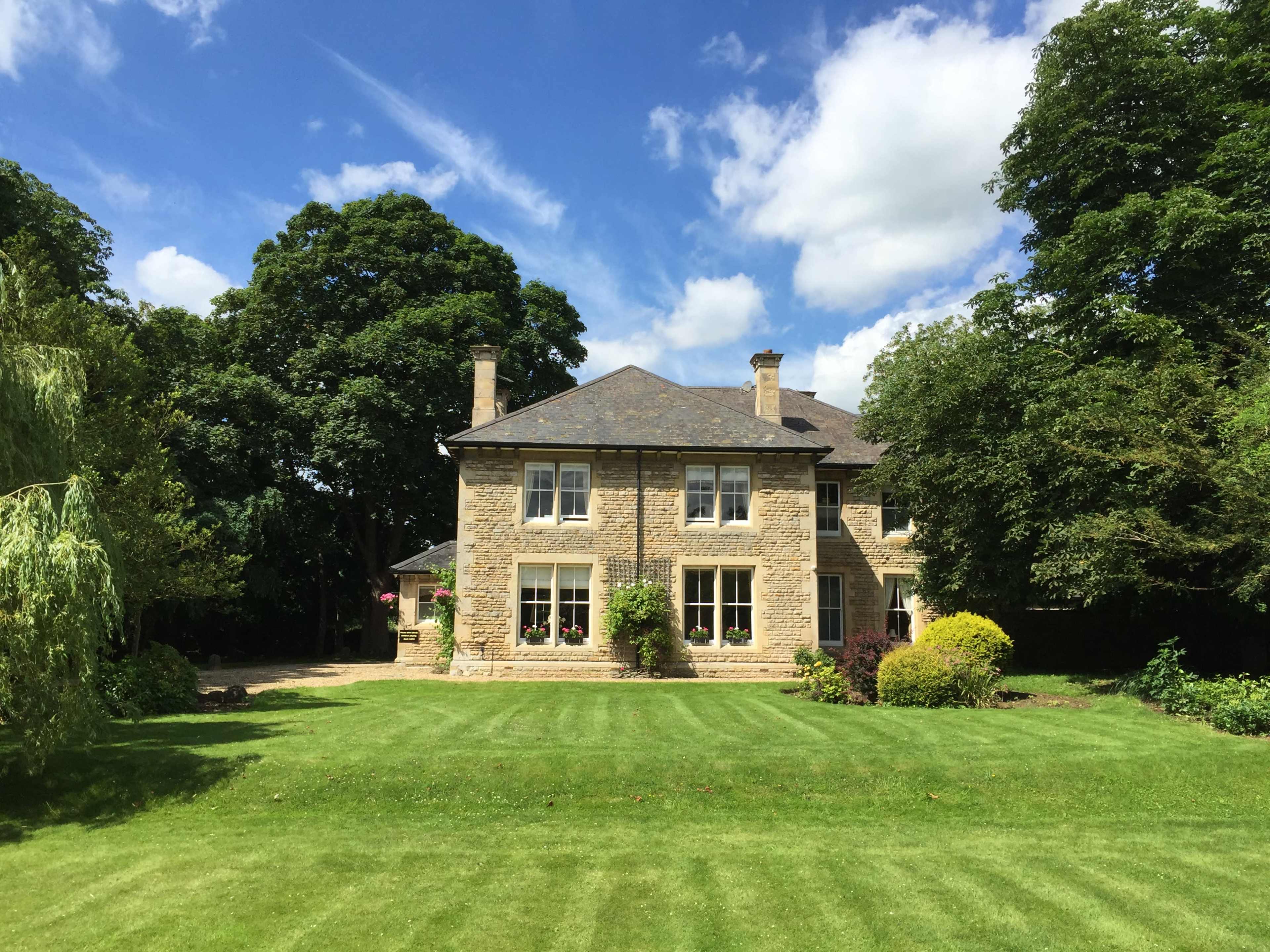 A large stone house with several windows is situated on a well-manicured lawn surrounded by trees under a partly cloudy sky.