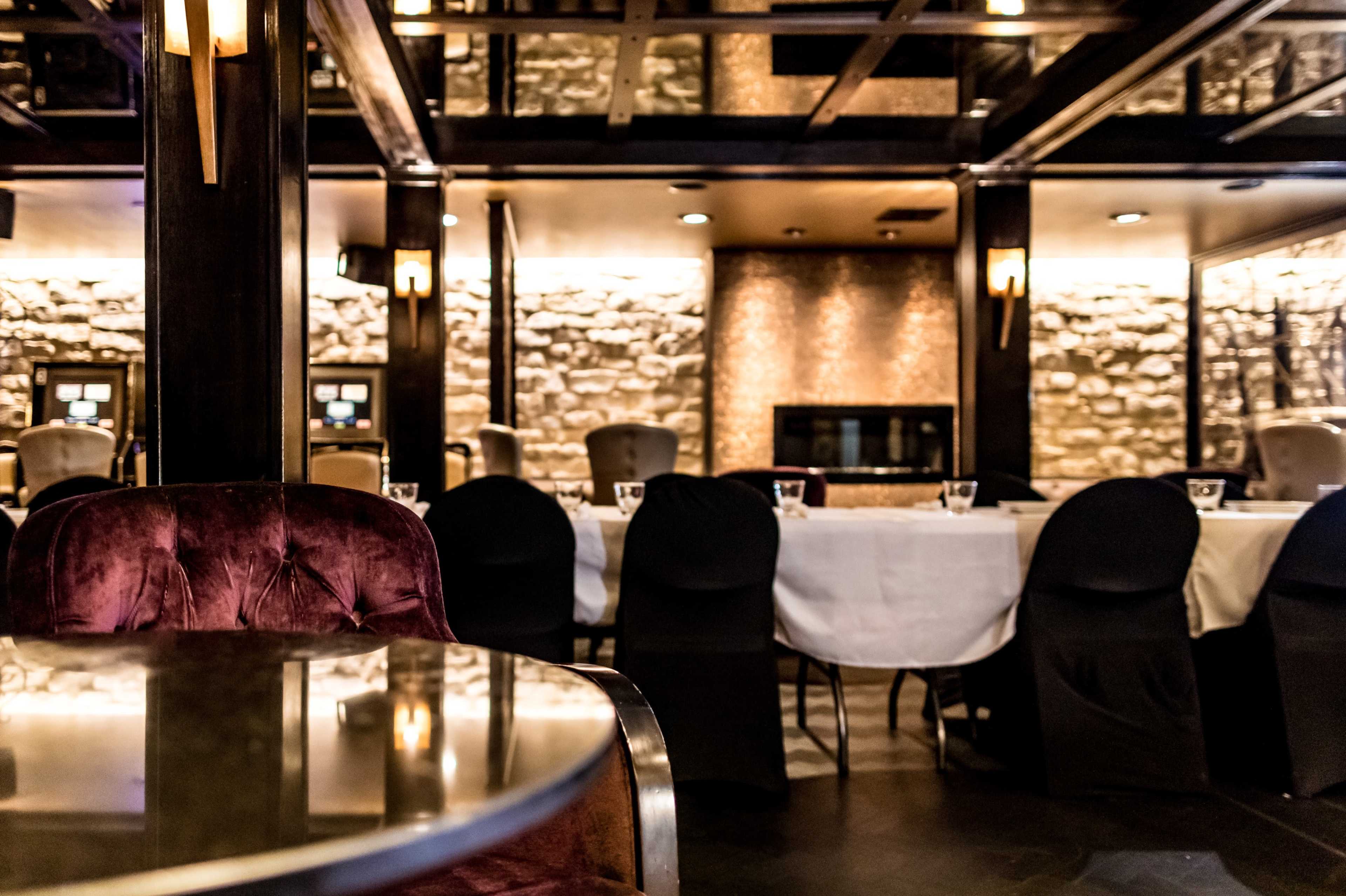 A dimly lit restaurant interior with a long white table set for dining, surrounded by dark chairs and stone walls.