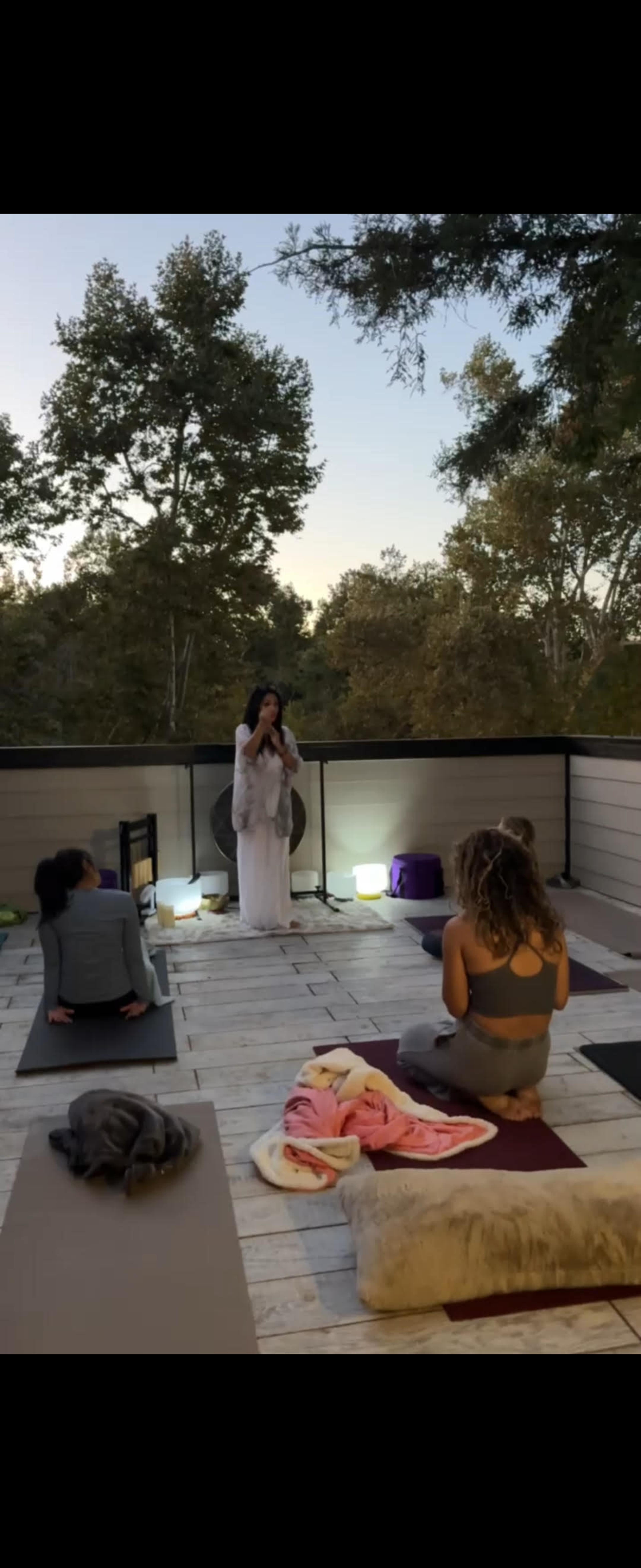 A group of individuals is practicing yoga on a wooden deck surrounded by trees during twilight.