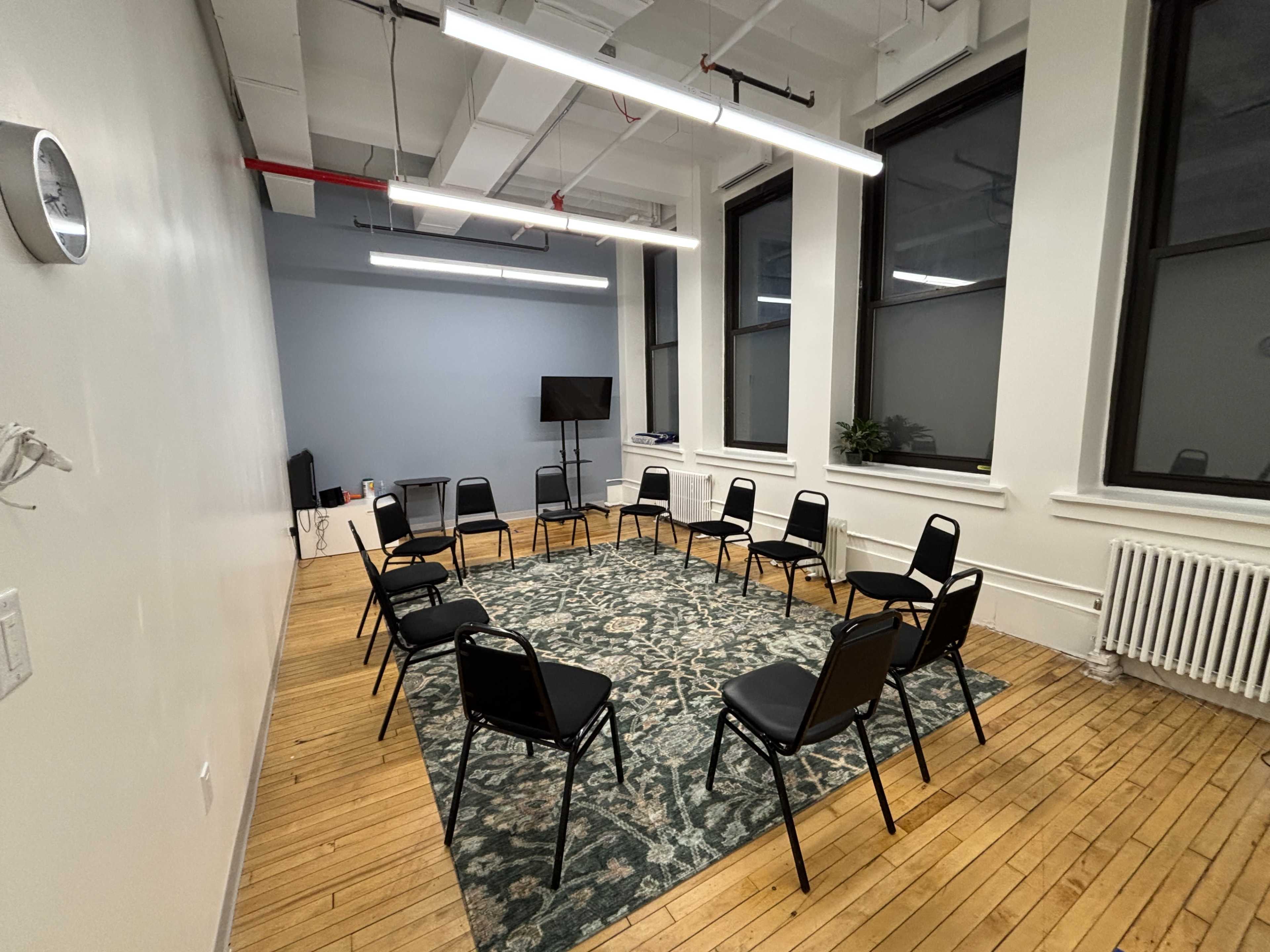 A meeting room features a circular arrangement of black chairs on a decorative rug, with a television and windowed walls visible in the background.