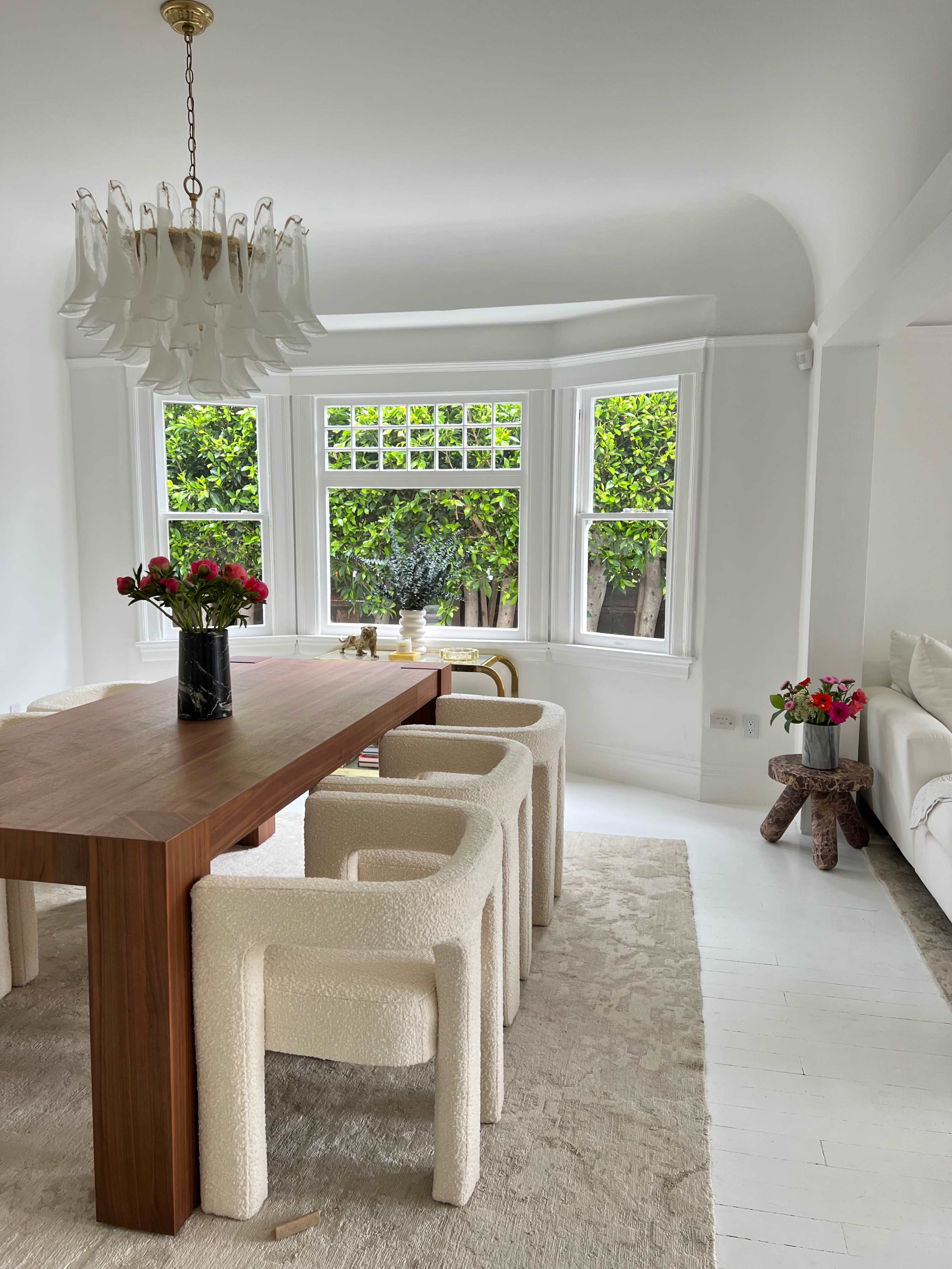 A dining area features a large wooden table surrounded by upholstered chairs, with a chandelier overhead and greenery visible through the windows.
