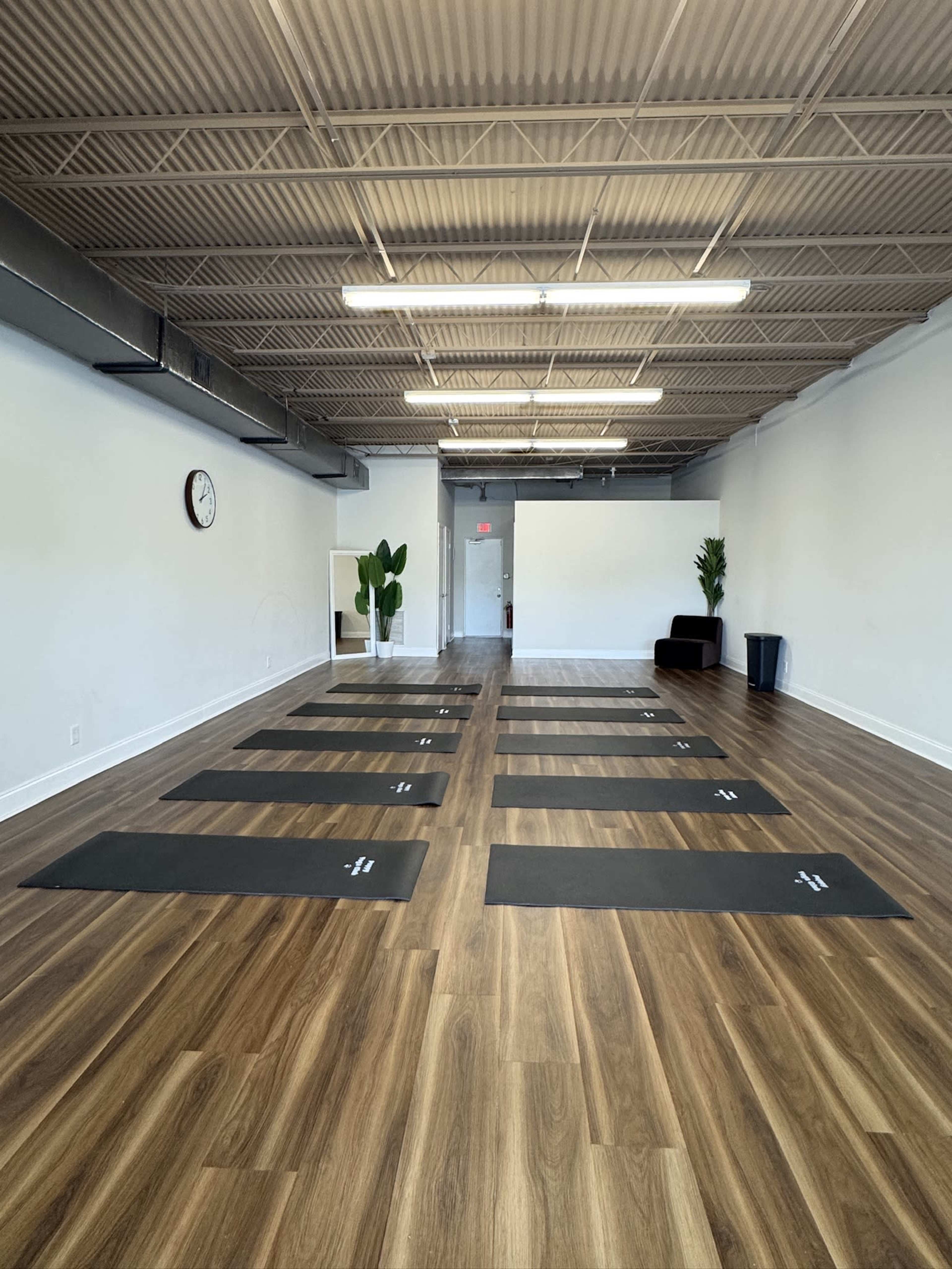 The image shows an empty yoga studio with several yoga mats arranged on a polished wooden floor, a clock on the wall, and a few potted plants in the corners.