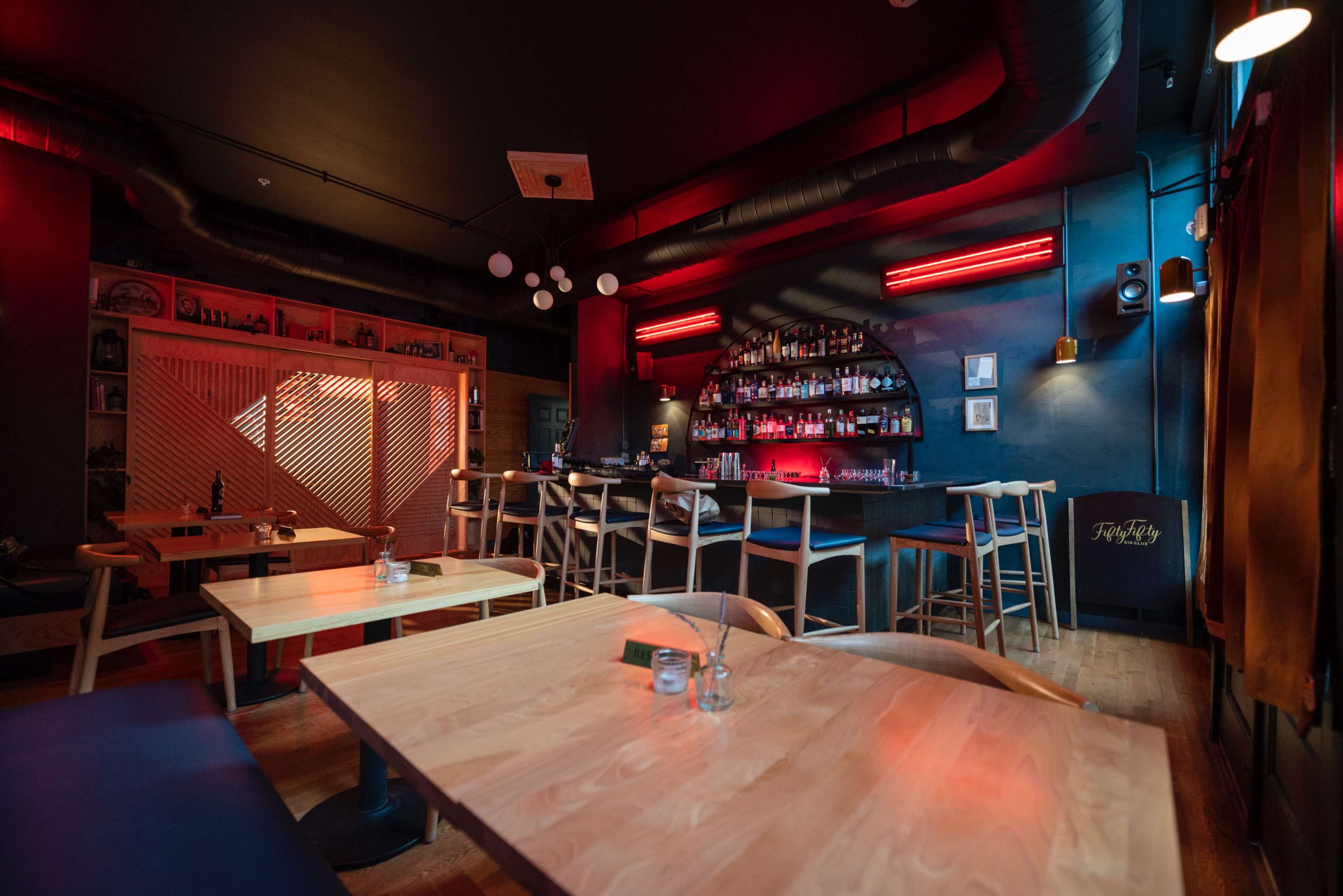 A modern bar interior features wooden tables and a well-stocked backlit bar against a dark blue wall, illuminated by red neon lights.