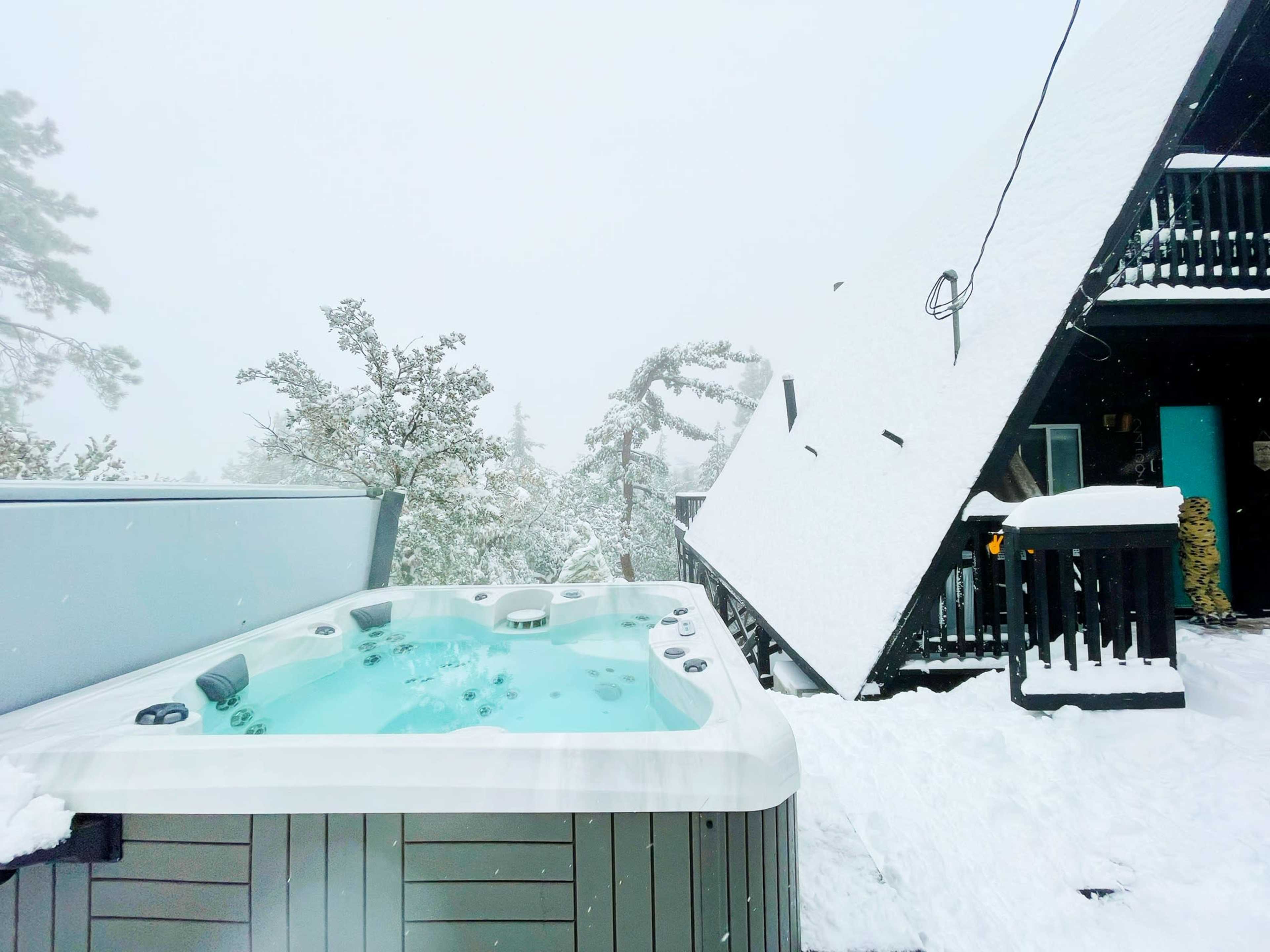 A hot tub sits next to a snow-covered cabin with a sloped roof in a snowy landscape.