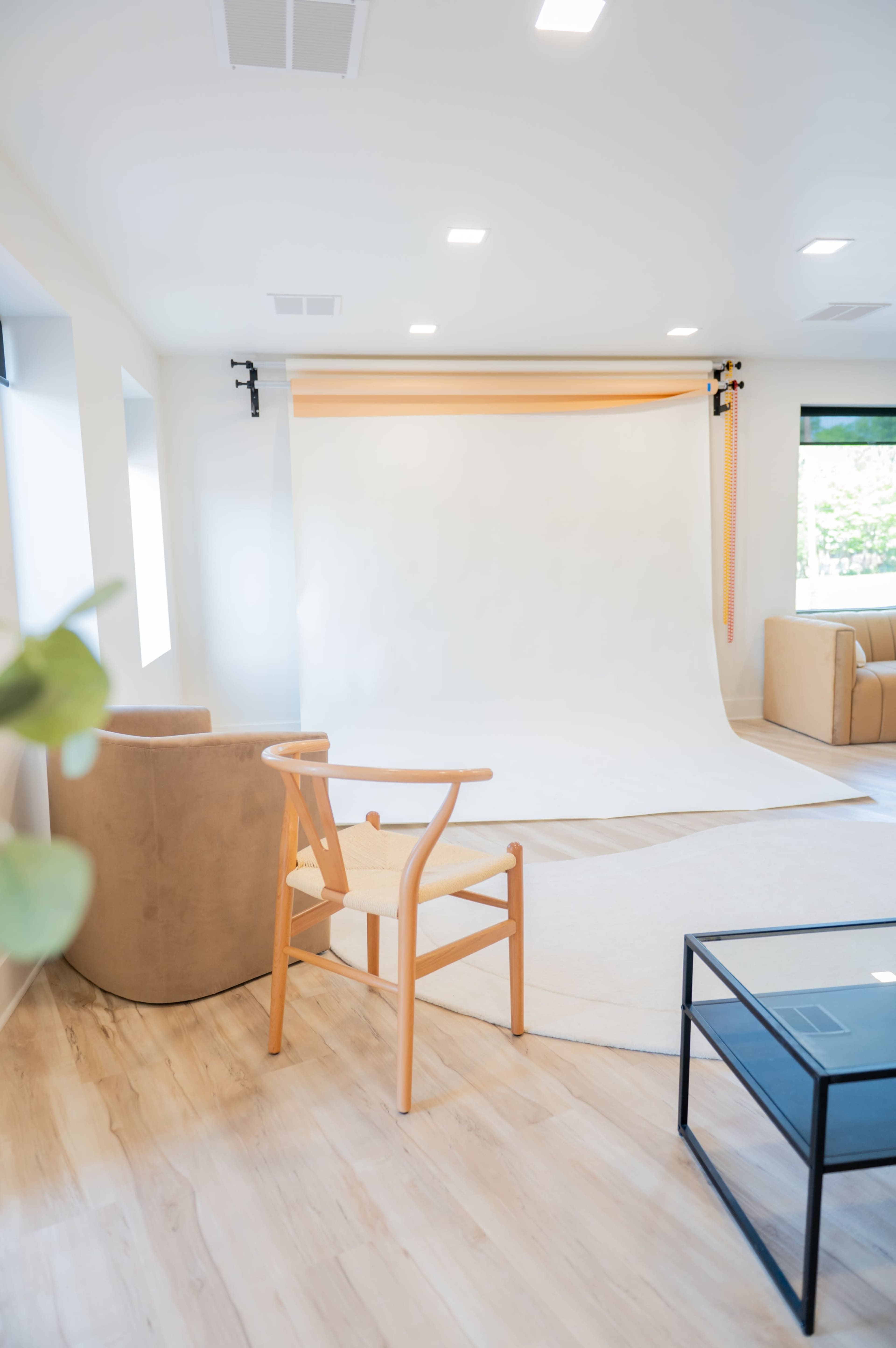 A minimalist photo studio featuring a white backdrop, a wooden chair, a couch, and a coffee table on a light wooden floor.