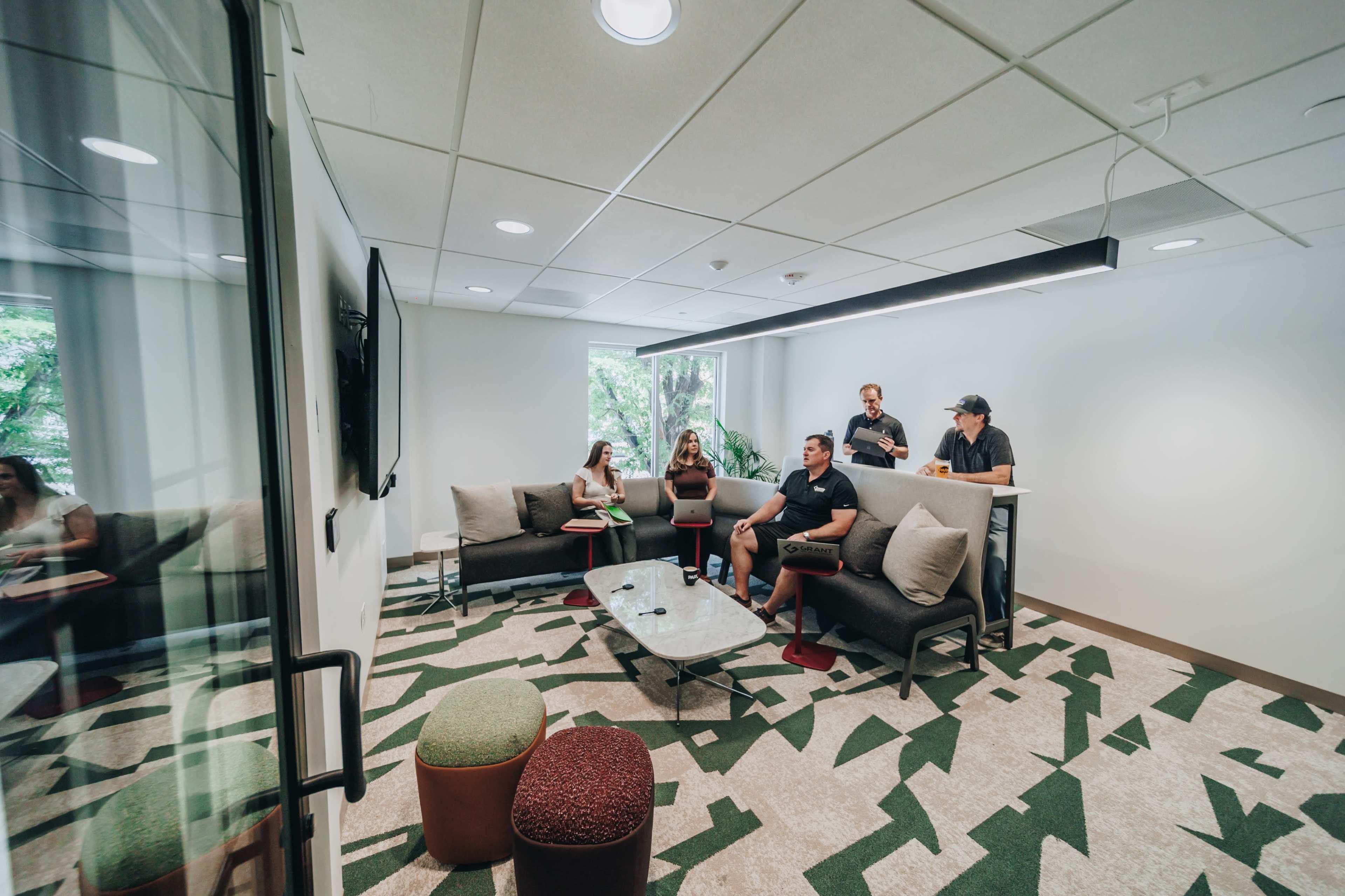 A group of five people is gathered in a modern meeting room with large windows, sitting on couches and a high table, while two individuals stand nearby.