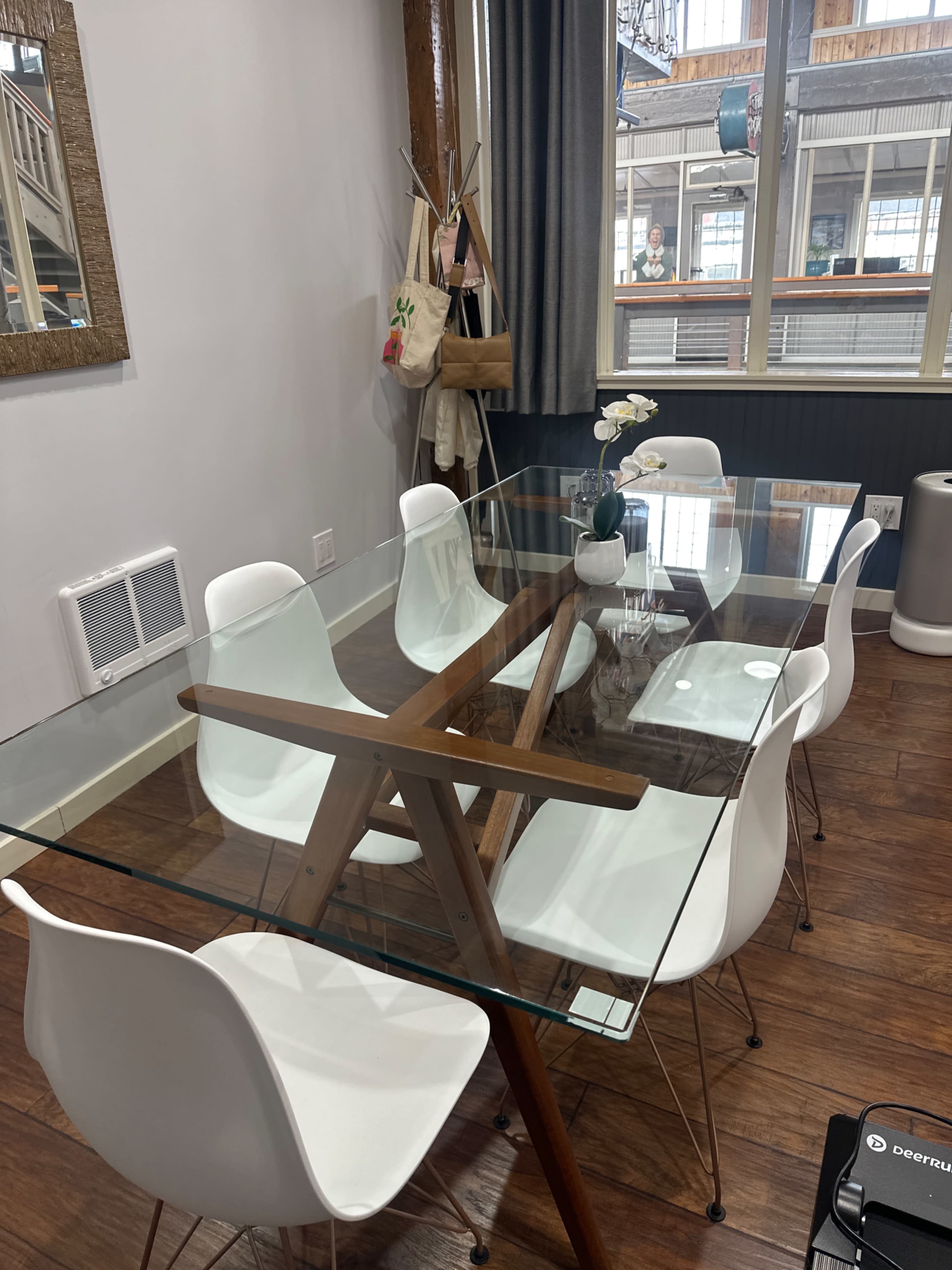 A glass dining table with wooden legs and white chairs is set in a well-lit room featuring large windows and light-colored walls.