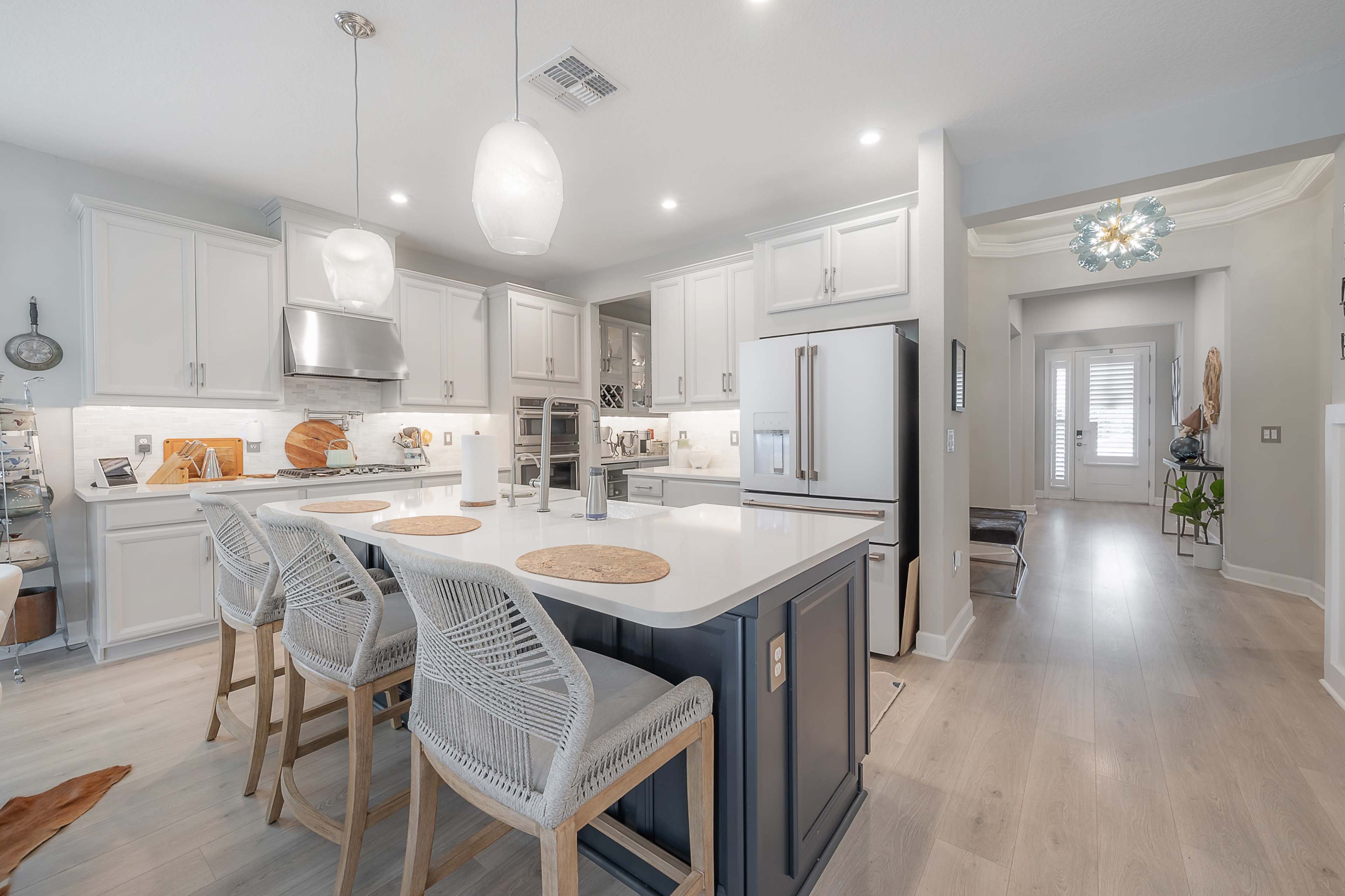 The image shows a modern kitchen with a large island, light-colored cabinets, and stainless steel appliances, transitioning into a hallway with a decorative light fixture and plants.
