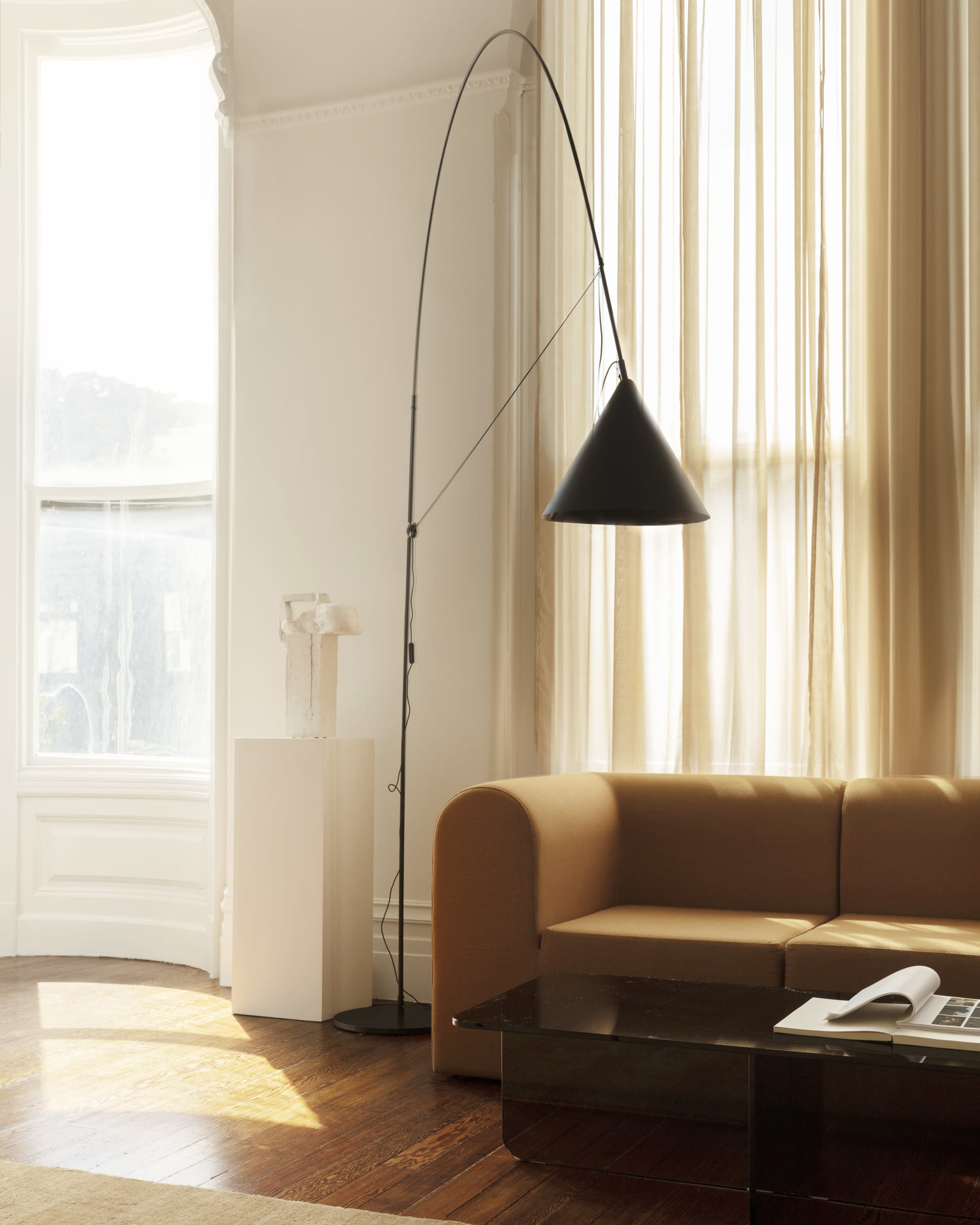 A modern living room featuring a curved floor lamp, a beige sofa, and a dark coffee table, with large windows letting in natural light.