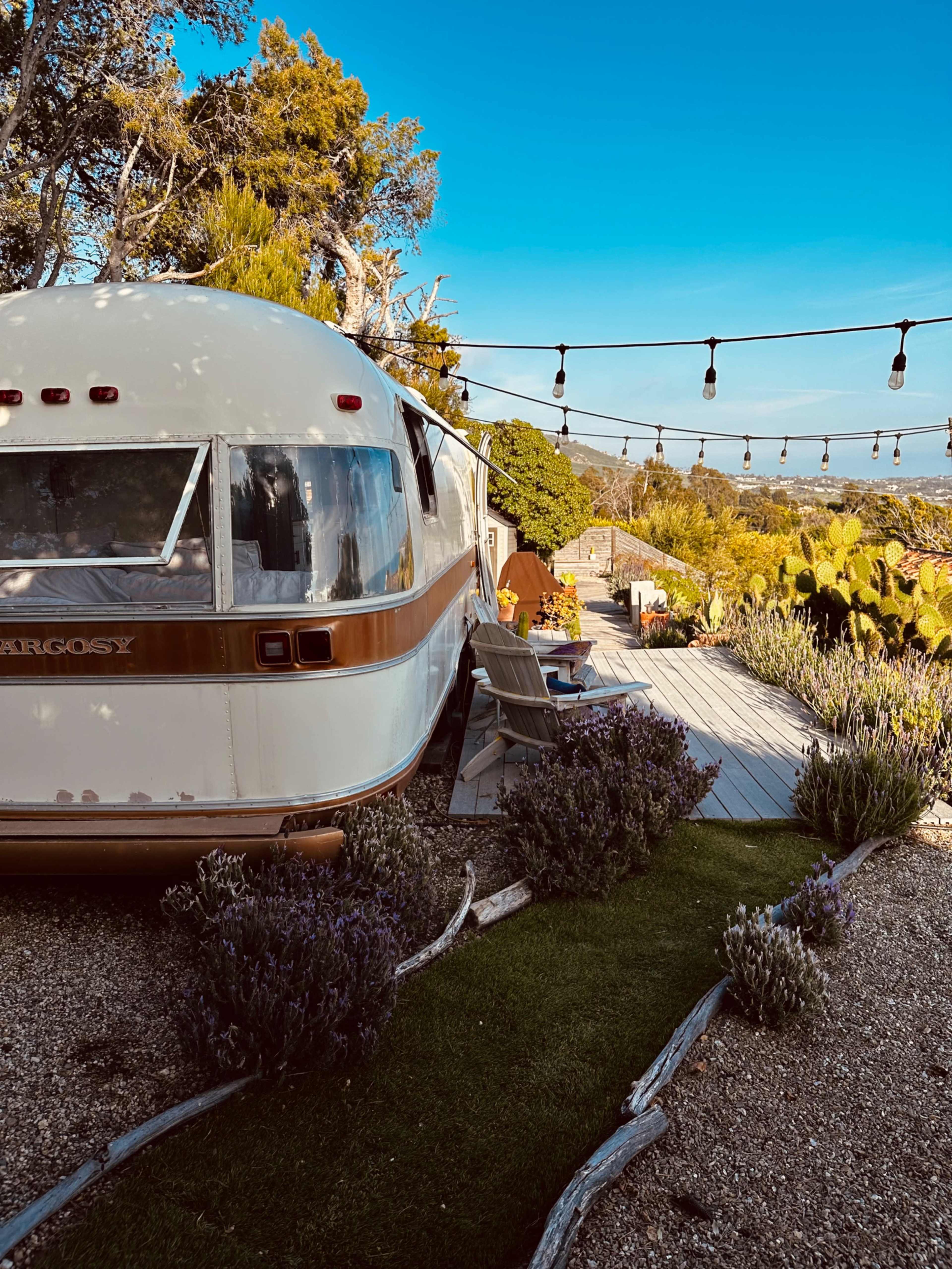 A vintage trailer is parked beside a garden with lavender and a path leading to outdoor seating under string lights.